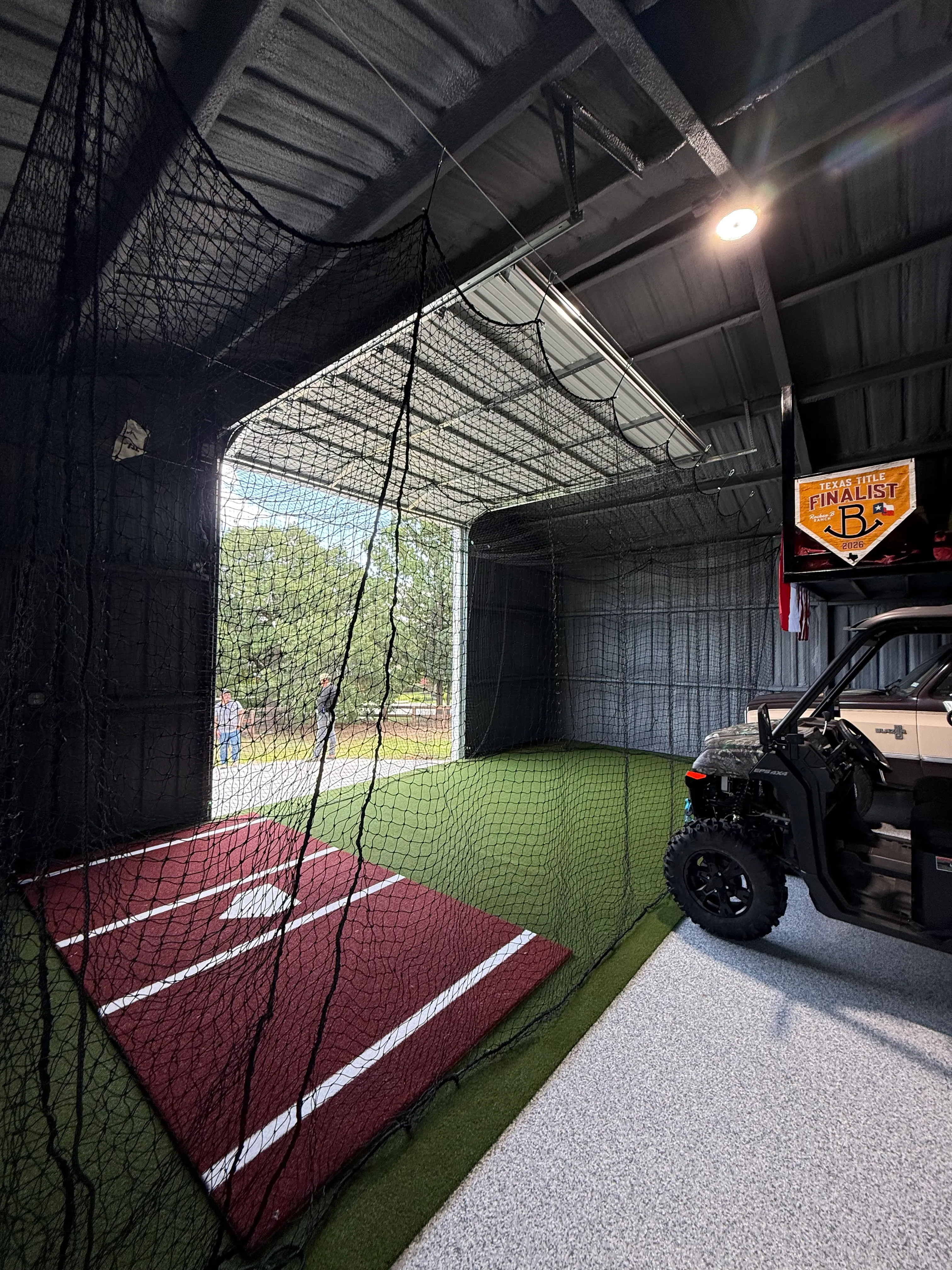 Batting cage netting and turf inside a working barn, with a UTV parked alongside near the open bay door, Argyle TX