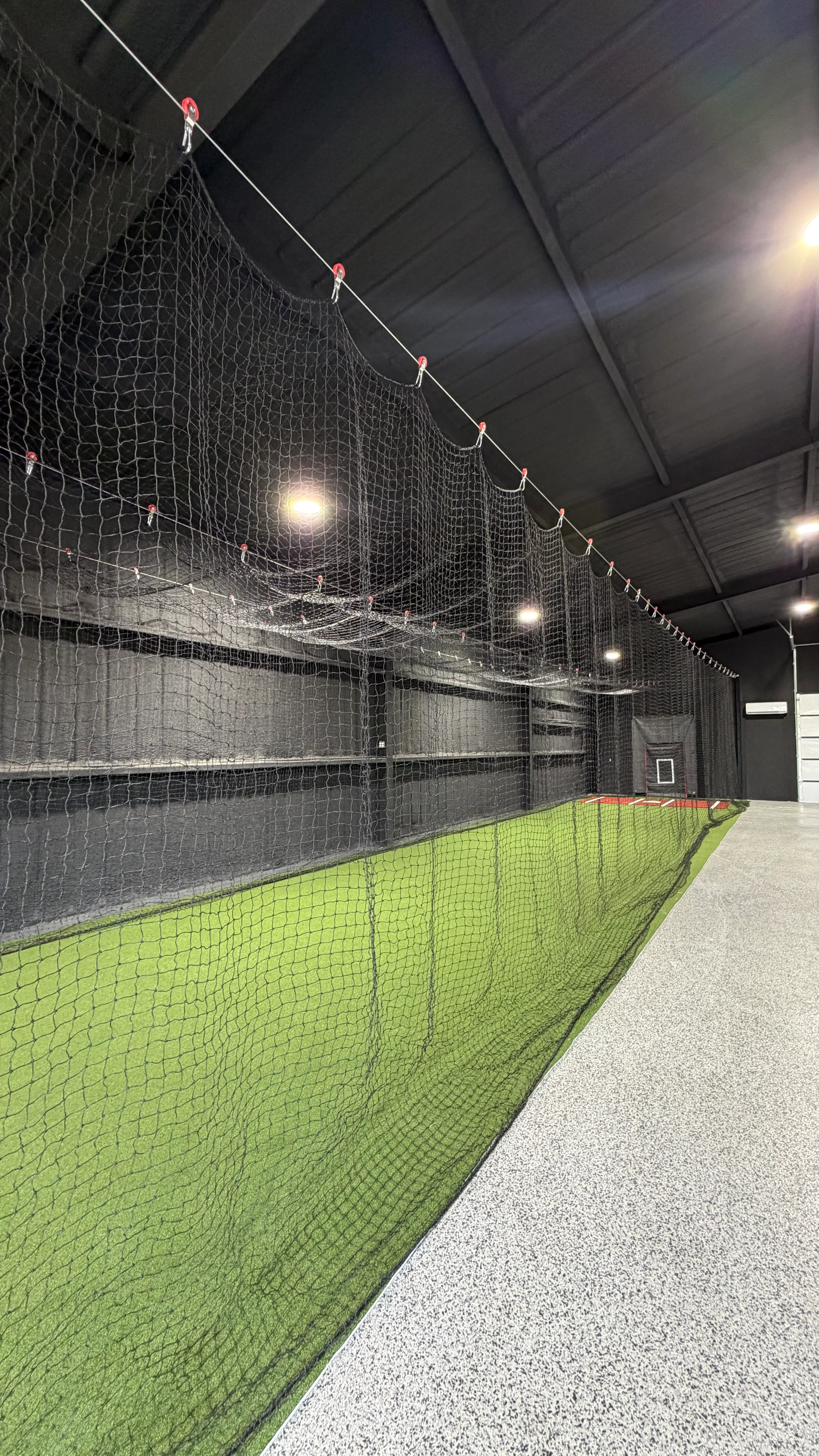 Vertical view down a long indoor batting cage lane with cable-hung netting and turf flooring