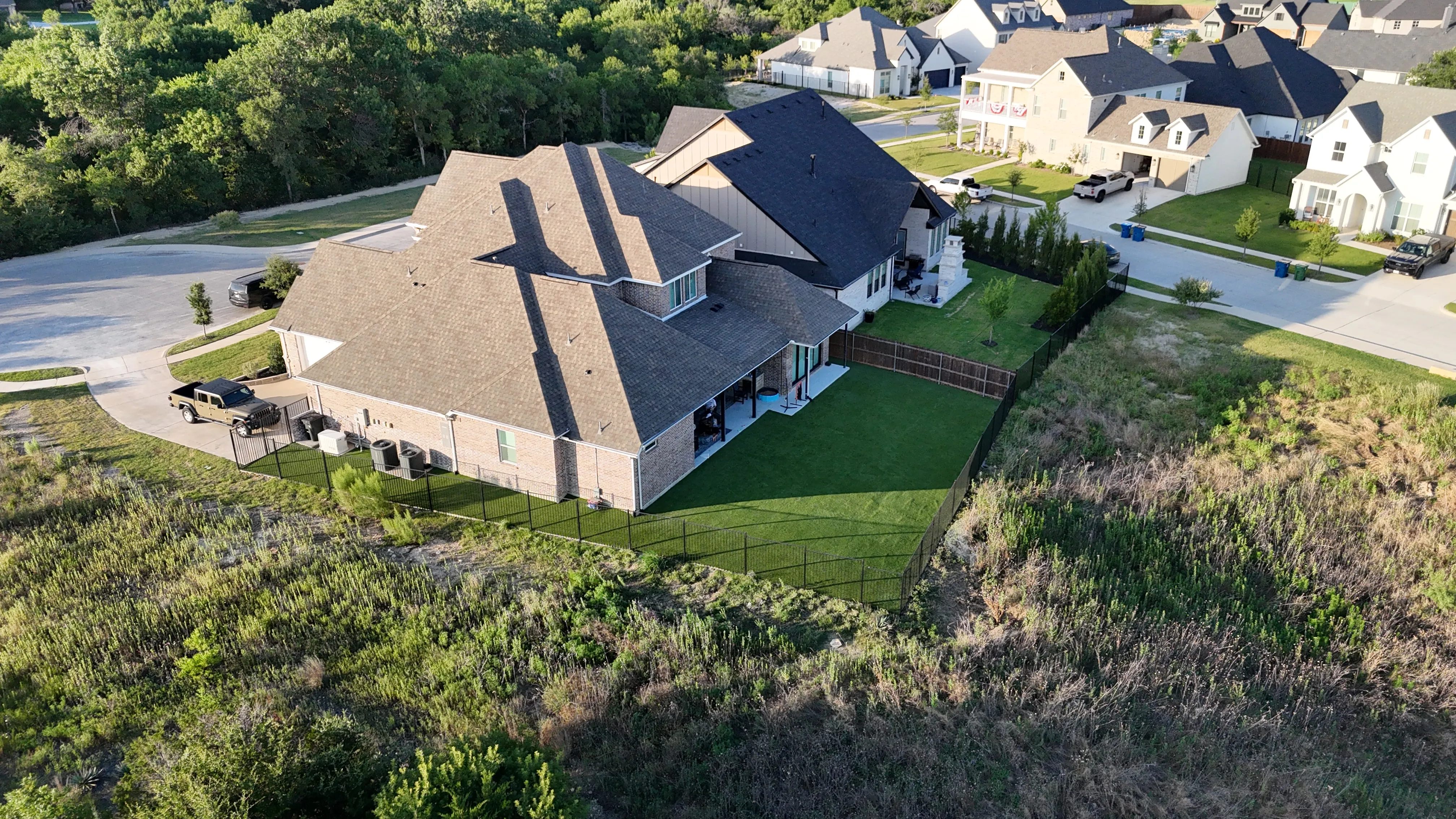 Aerial of a pie-shaped backyard with artificial turf lawn and outdoor stone fireplace bordering a wooded lot in DFW