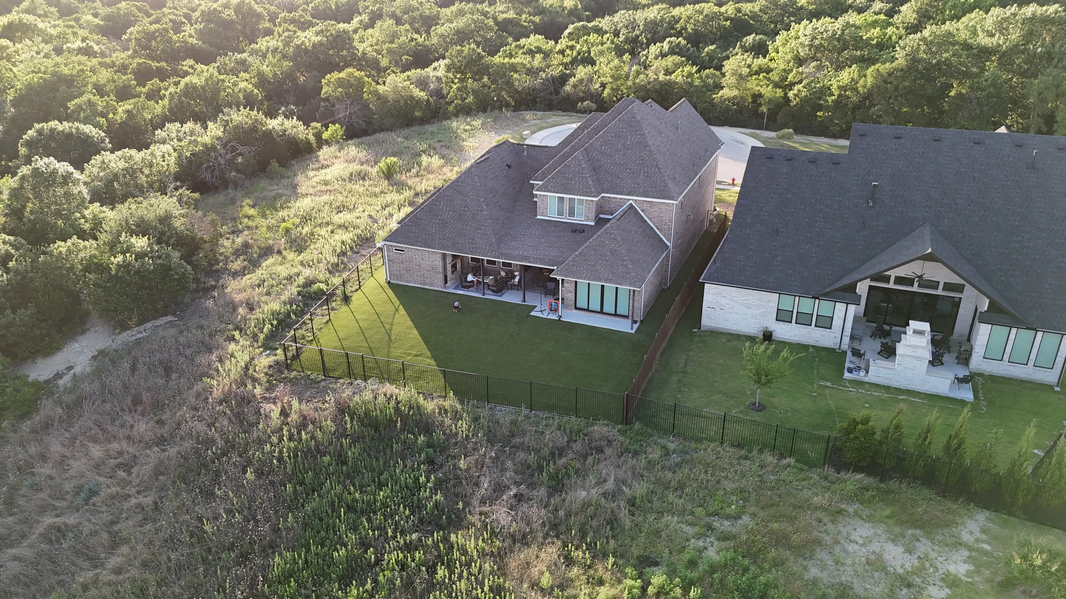 Aerial of two adjoining DFW backyards with artificial turf lawns bordering a wooded lot, families relaxing on covered patios