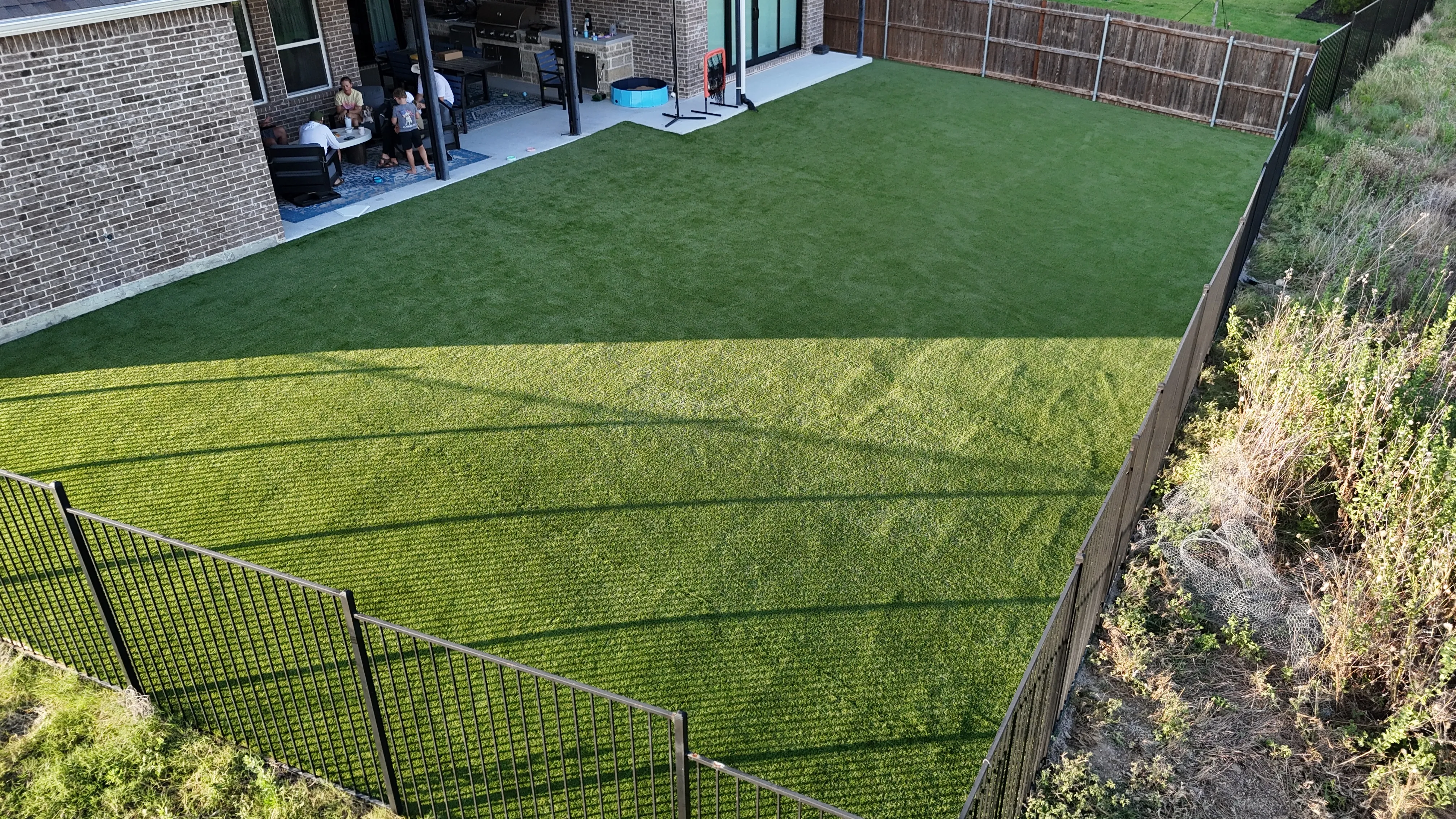 Close aerial detail of artificial turf blades and lawn texture with a family gathered on the covered patio in DFW