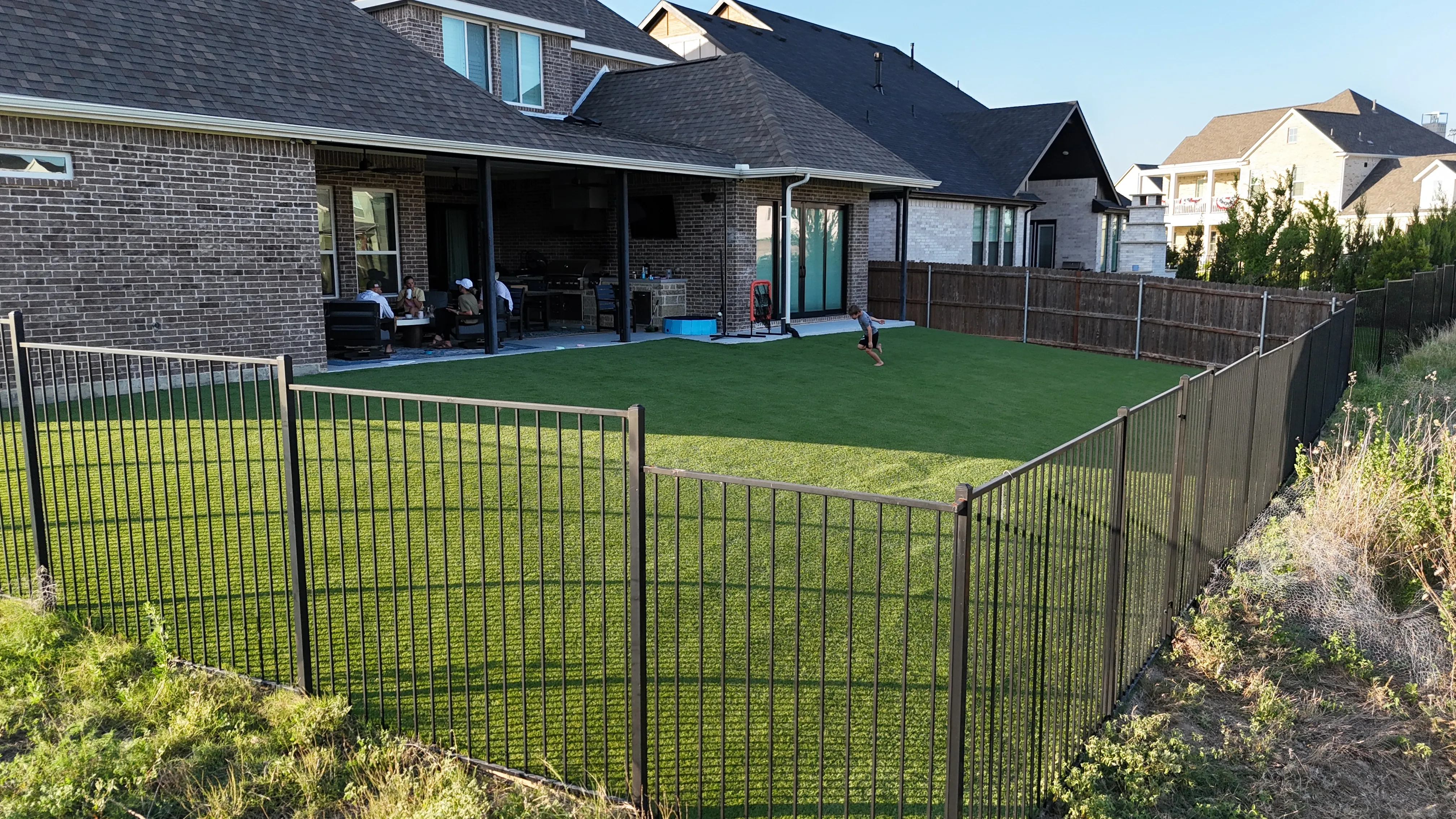Kid running across an artificial turf backyard while family relaxes on the covered patio in DFW