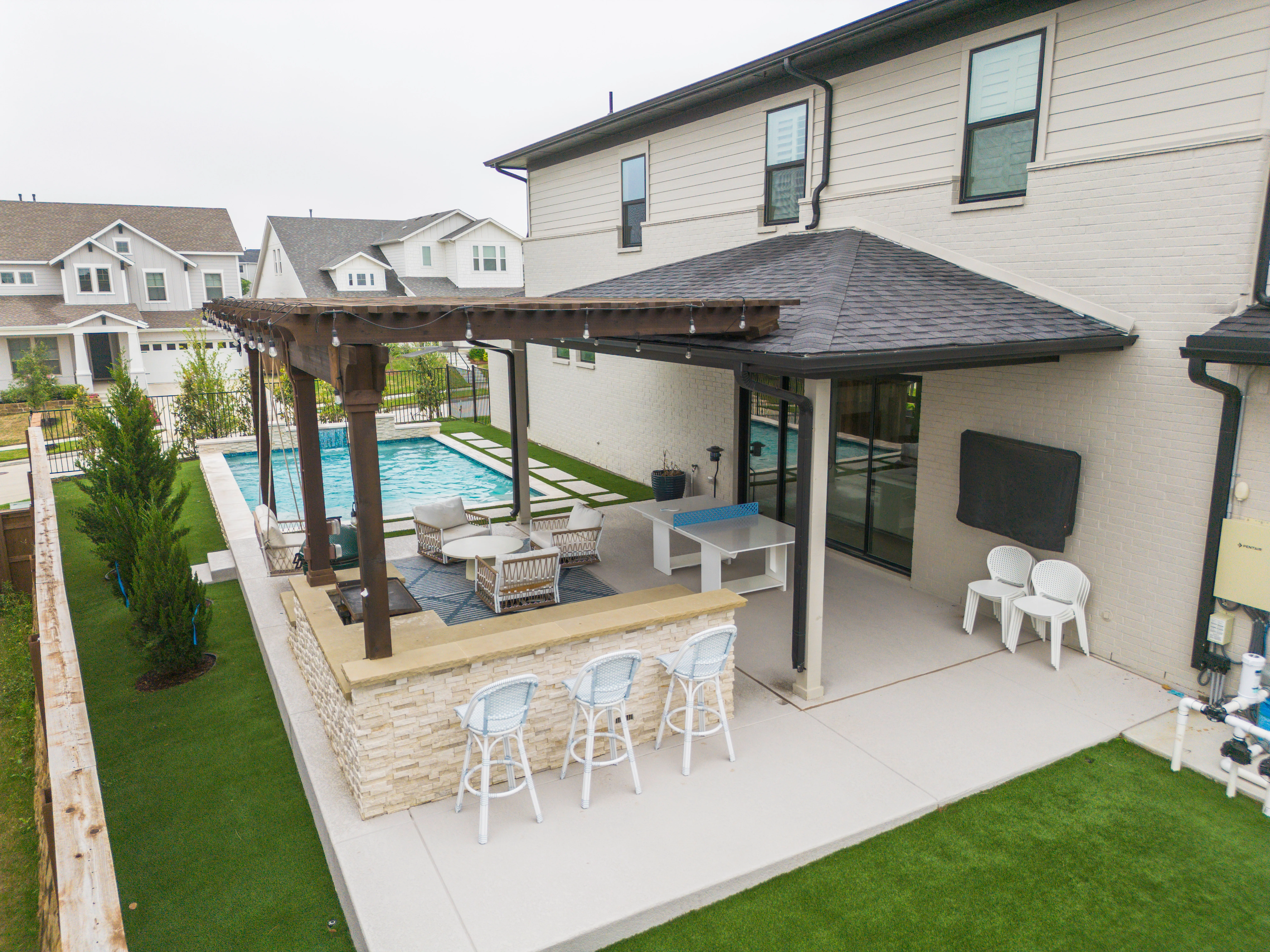 Elevated view of a DFW backyard with pool, artificial turf border, and a pergola-covered outdoor kitchen with bar seating and a ping pong table