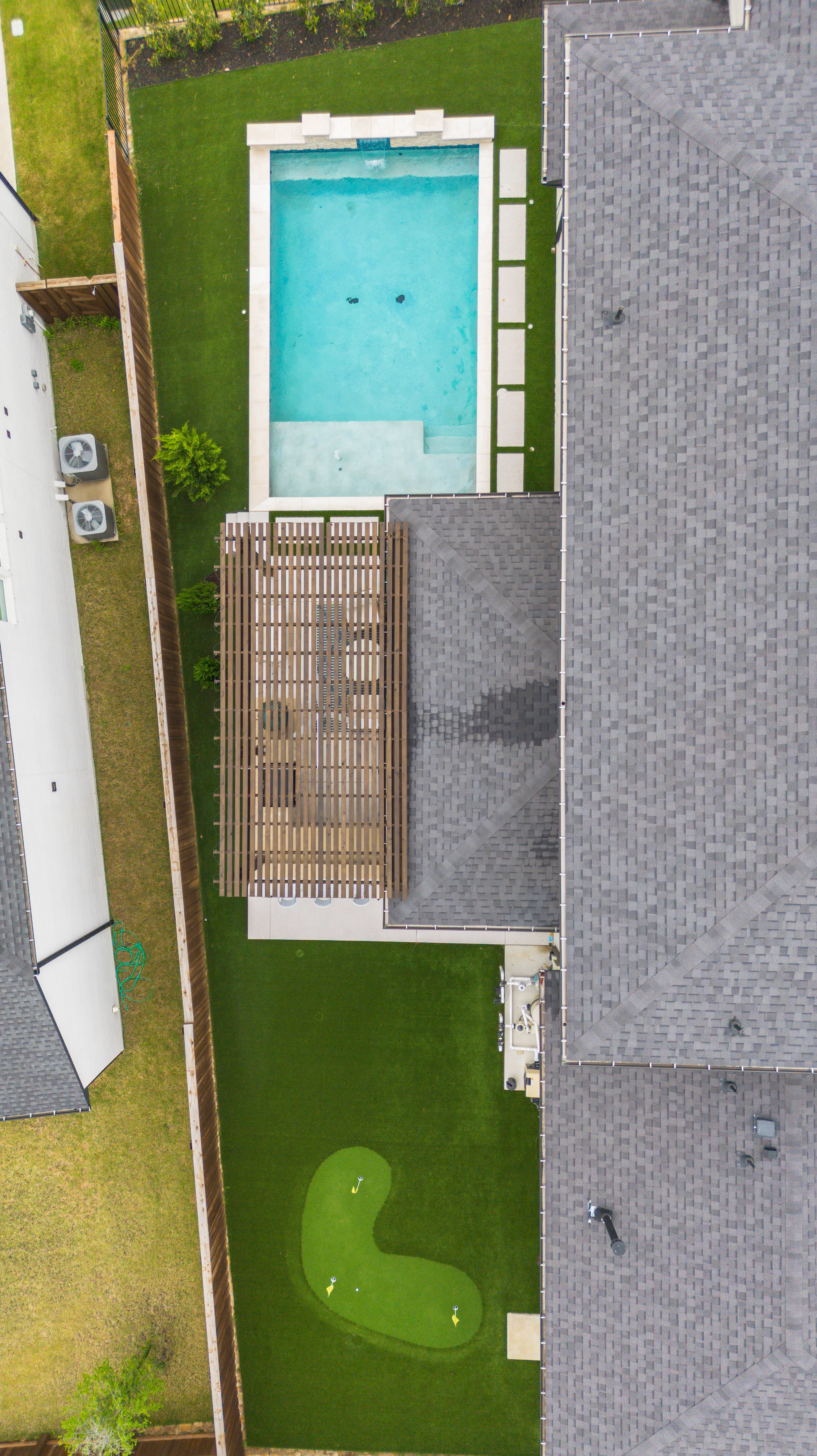 Top-down aerial of a DFW backyard pool and slatted pergola roof beside a curved putting green with three flags