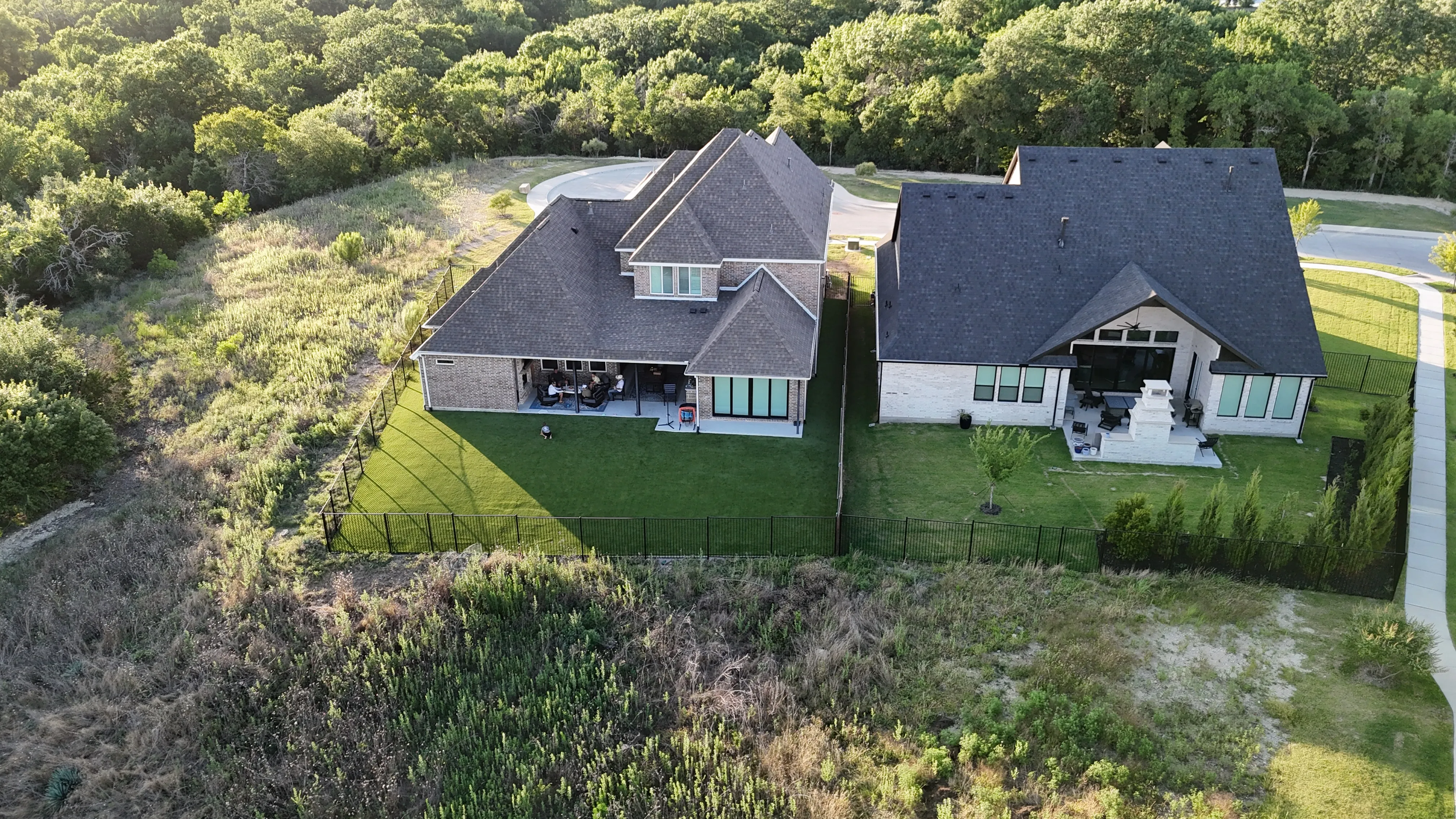 Aerial of two neighboring wooded-lot homes each with artificial turf backyards bordered by iron fencing