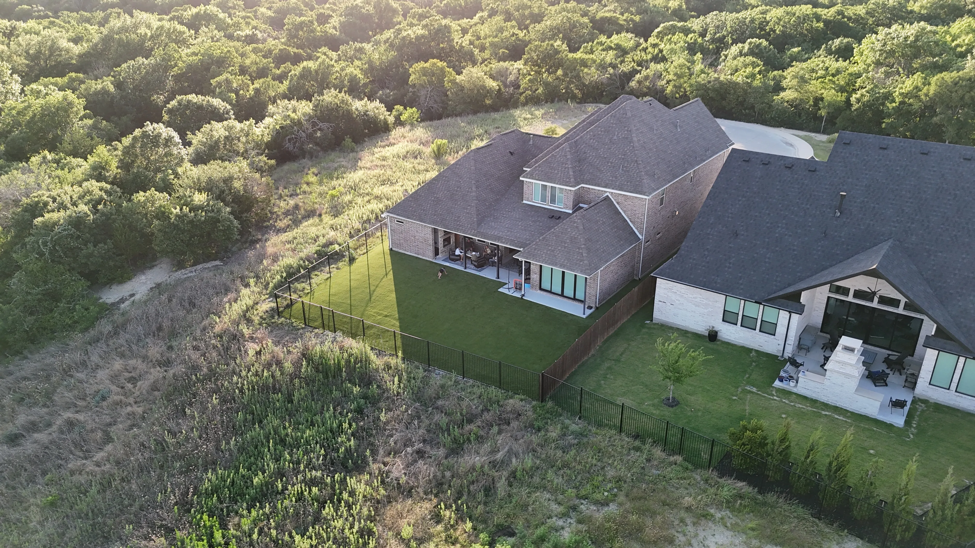 Aerial of a turf backyard with tall sports netting along one side and a dog on the lawn, beside a stone patio with an outdoor fireplace
