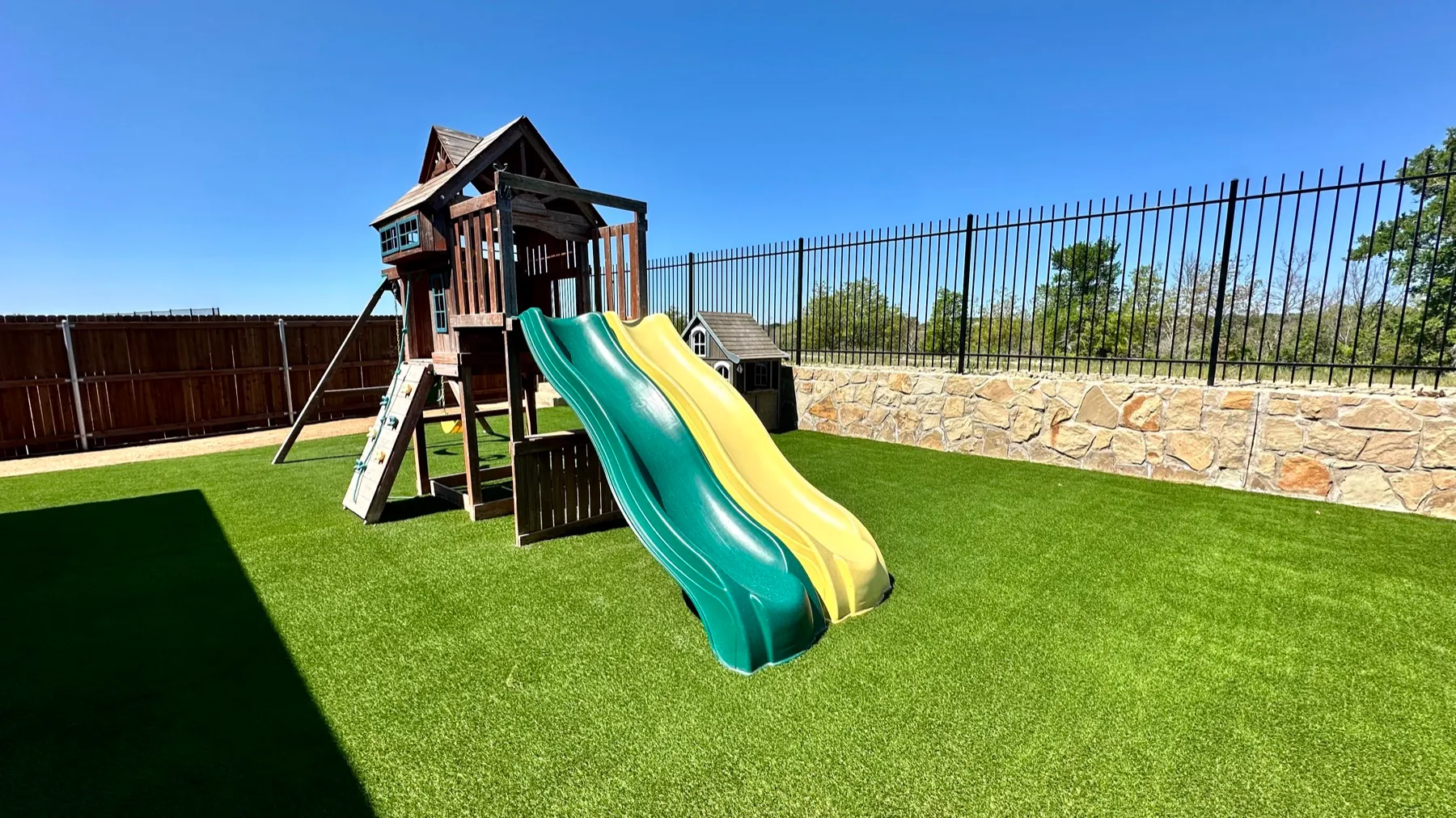 Backyard playset with double slide on turf beside stone wall