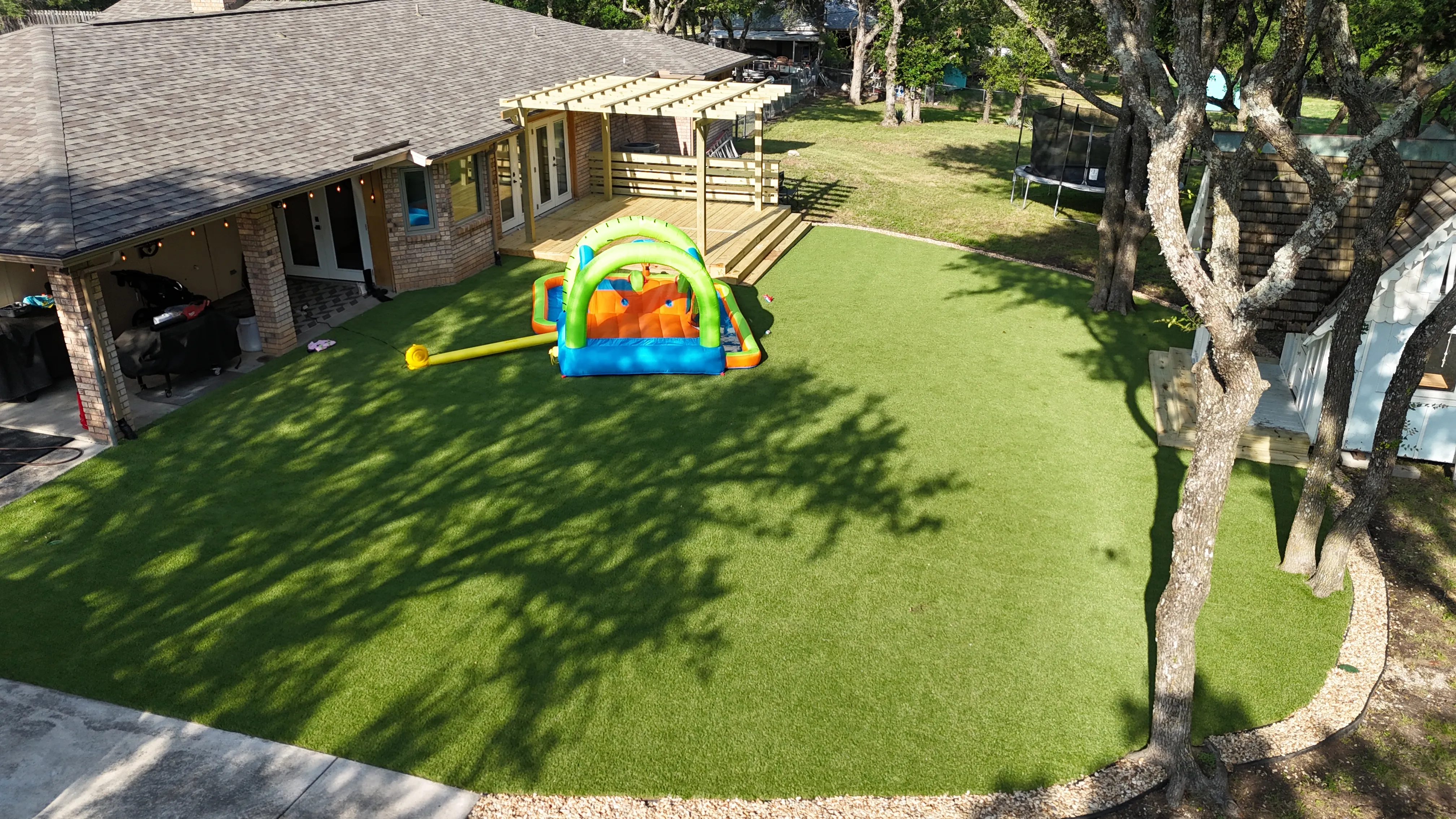 Aerial curved backyard artificial turf lawn under mature oak trees with inflatable water slide, DFW