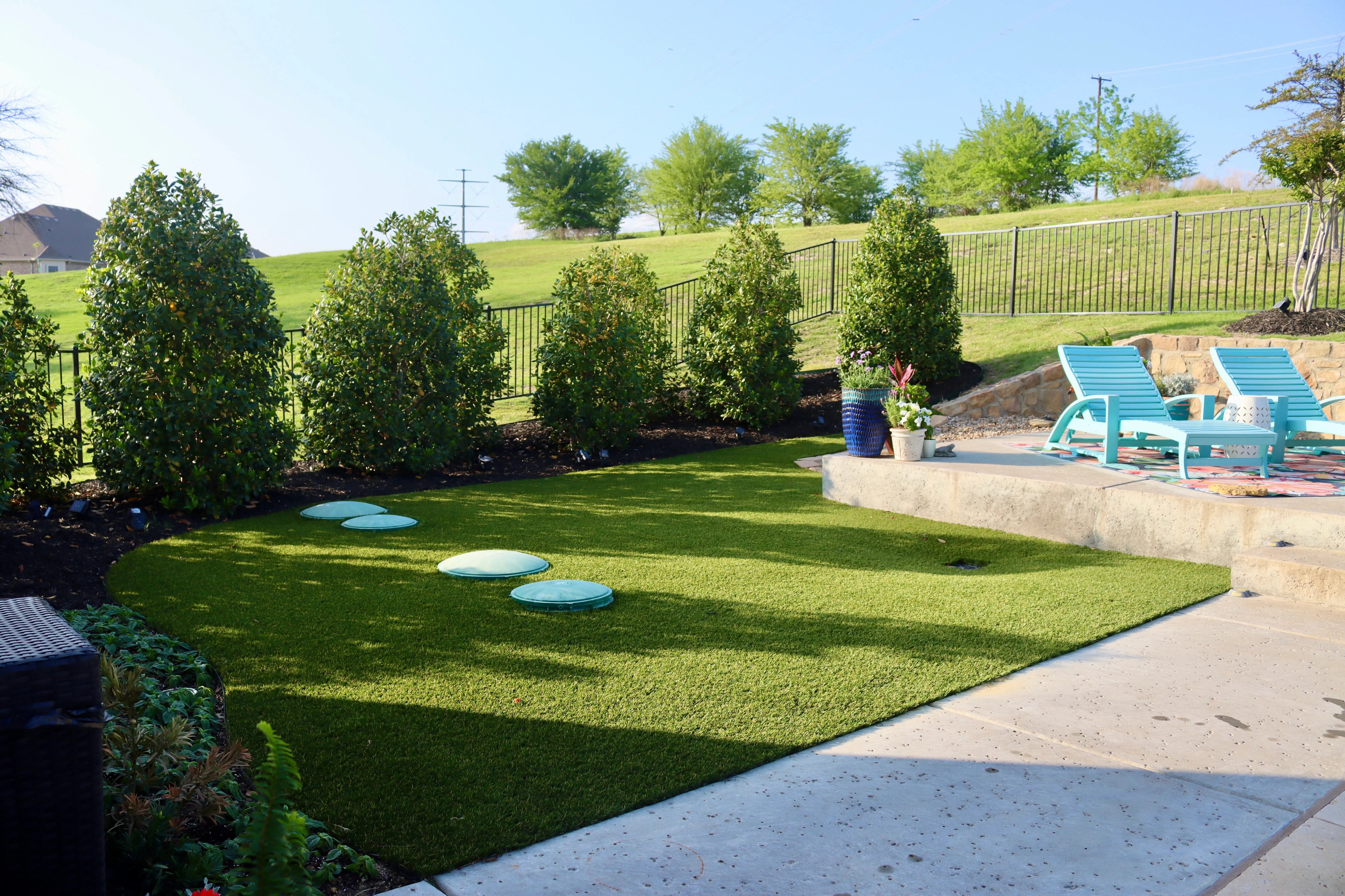 Curved artificial turf lawn bordered by clipped hedges with raised stone patio and lounge chairs in DFW