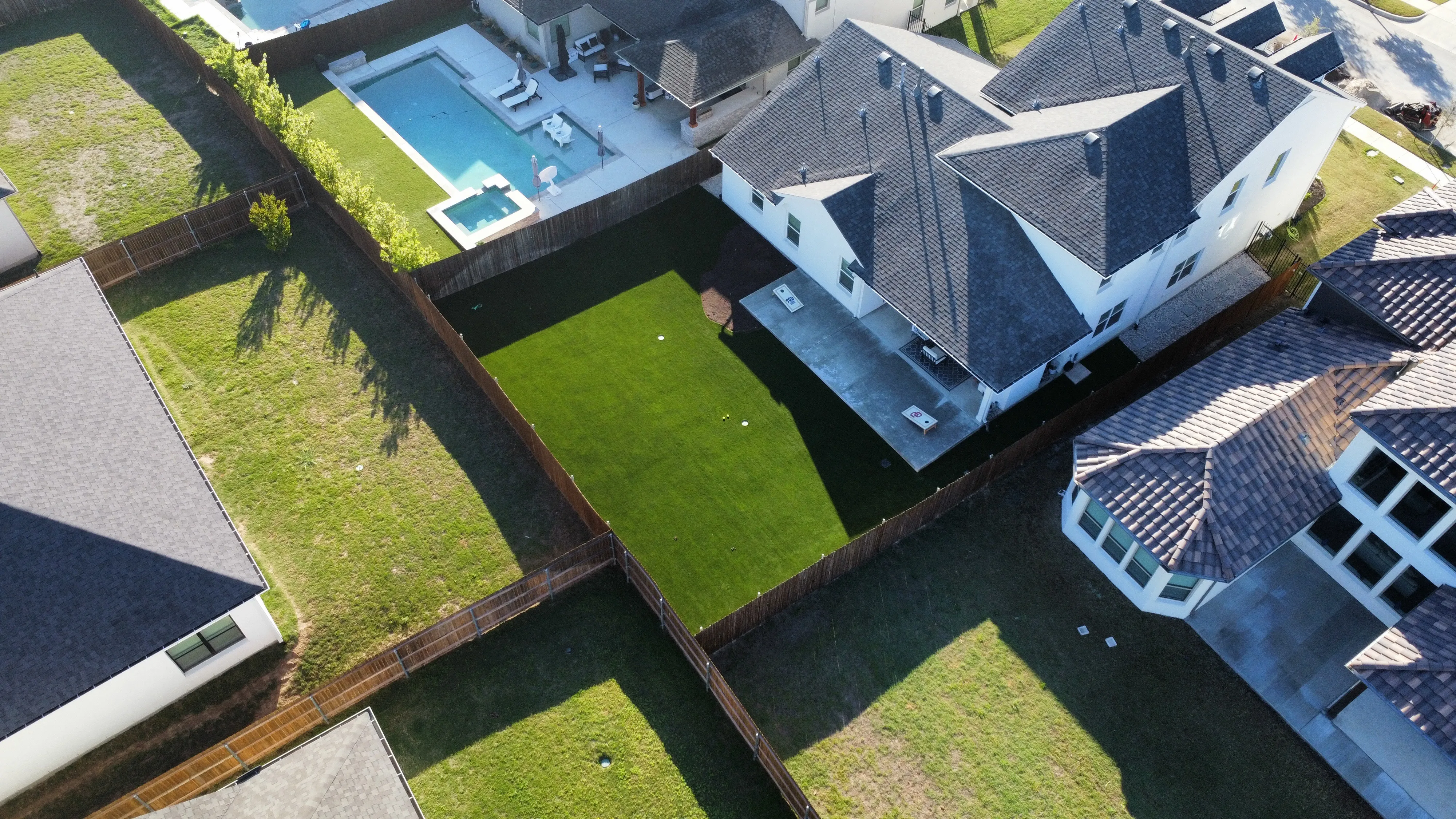Aerial backyard with artificial turf, cornhole boards, and a neighboring pool, green turf contrasting with dormant natural grass lots in DFW