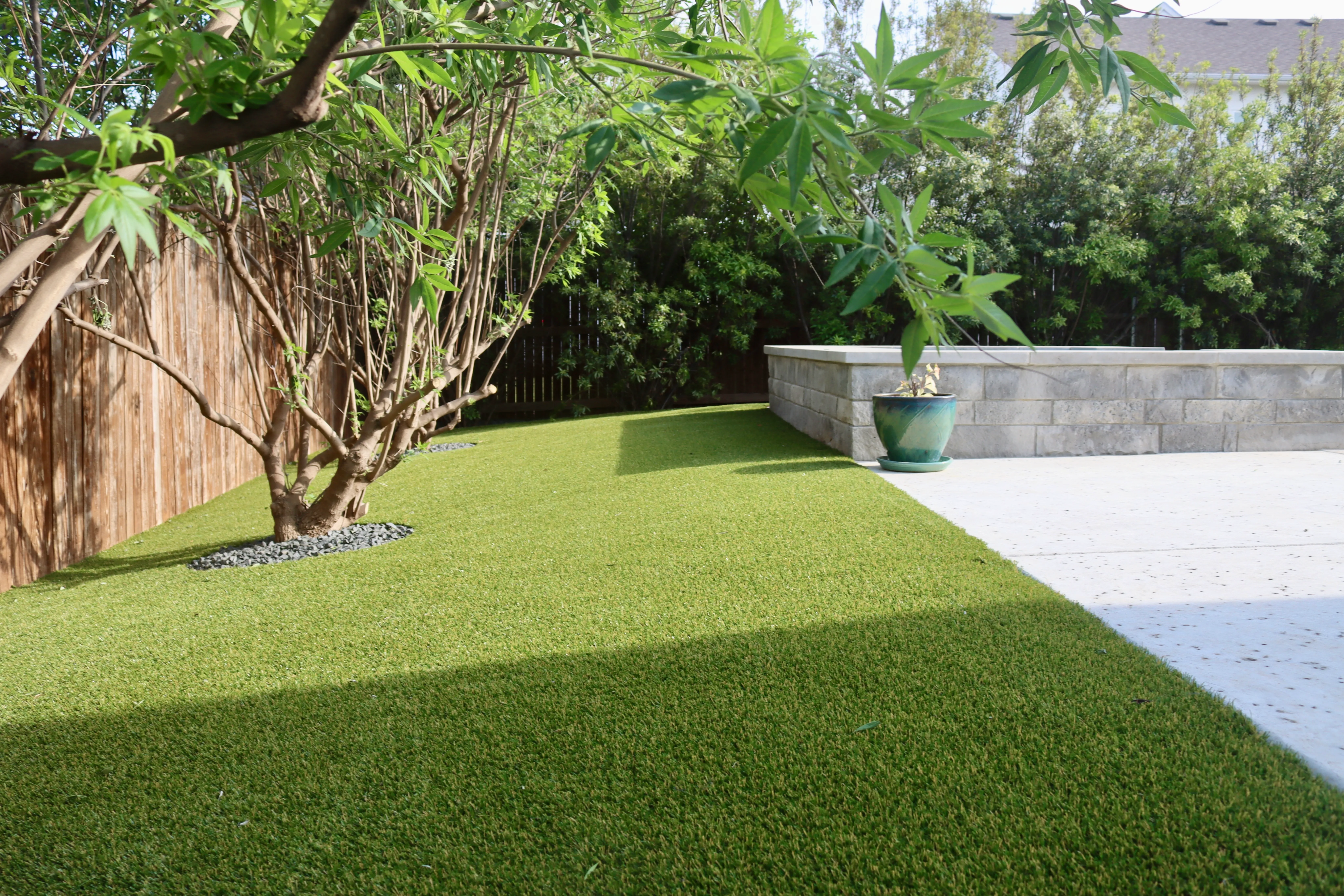 Artificial turf lawn beside a raised stone planter wall with a mature landscaping tree in a DFW backyard