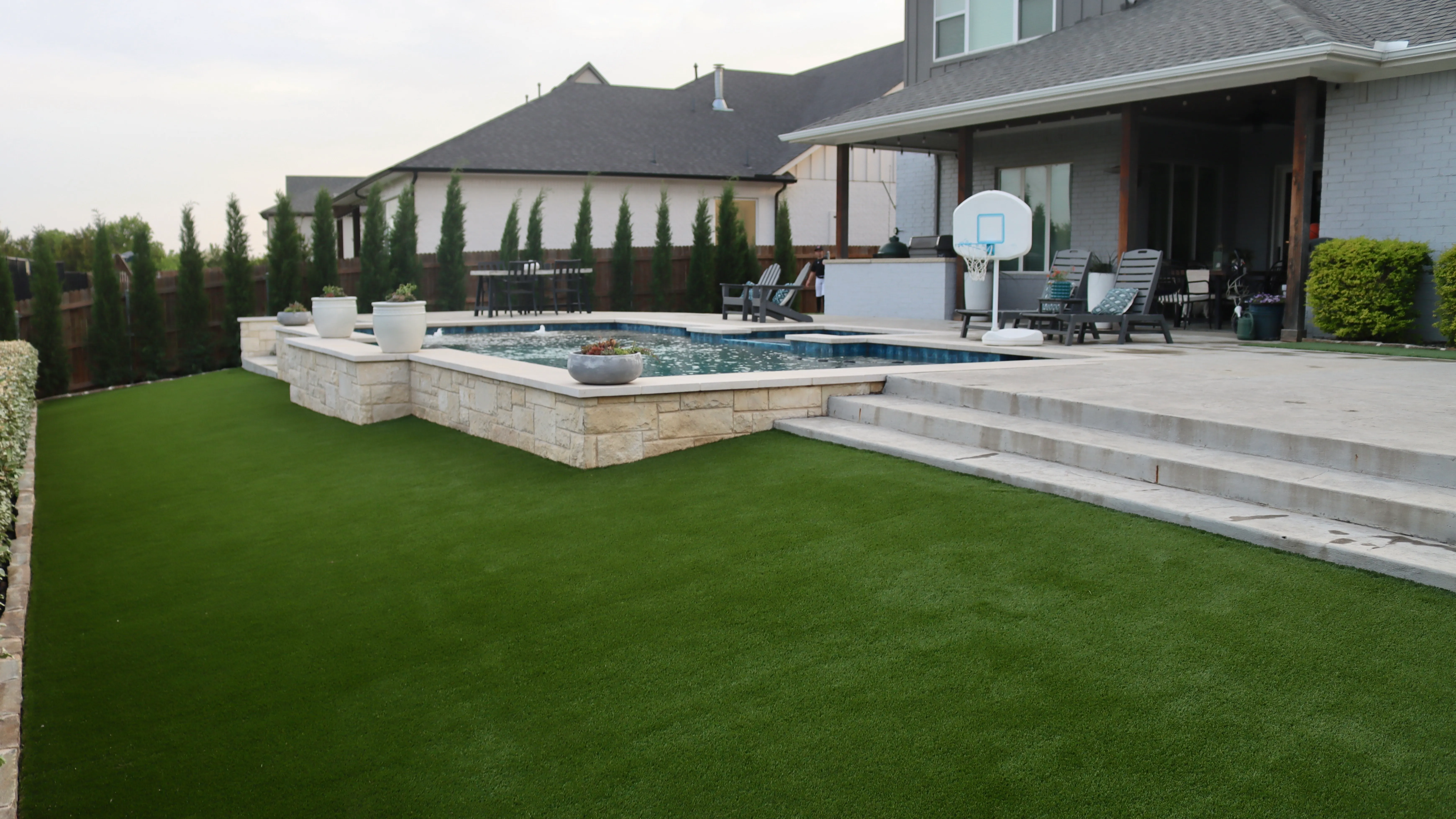 Elevated backyard pool terrace with stone retaining wall, cypress trees, basketball hoop, and artificial turf lawn in DFW