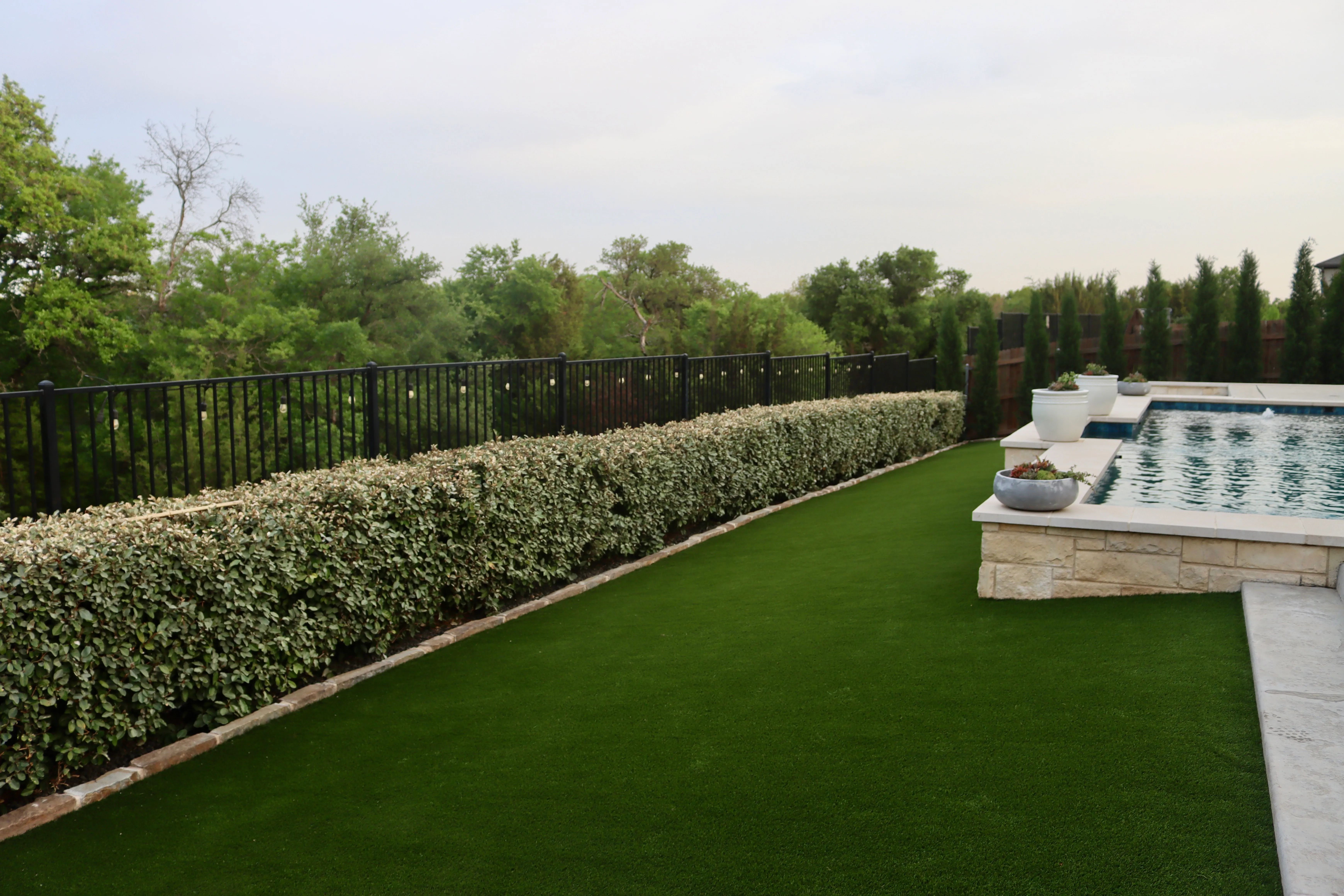 Wide low-angle view of an elevated stone pool wall bordered by a cypress hedge and artificial turf strip in DFW