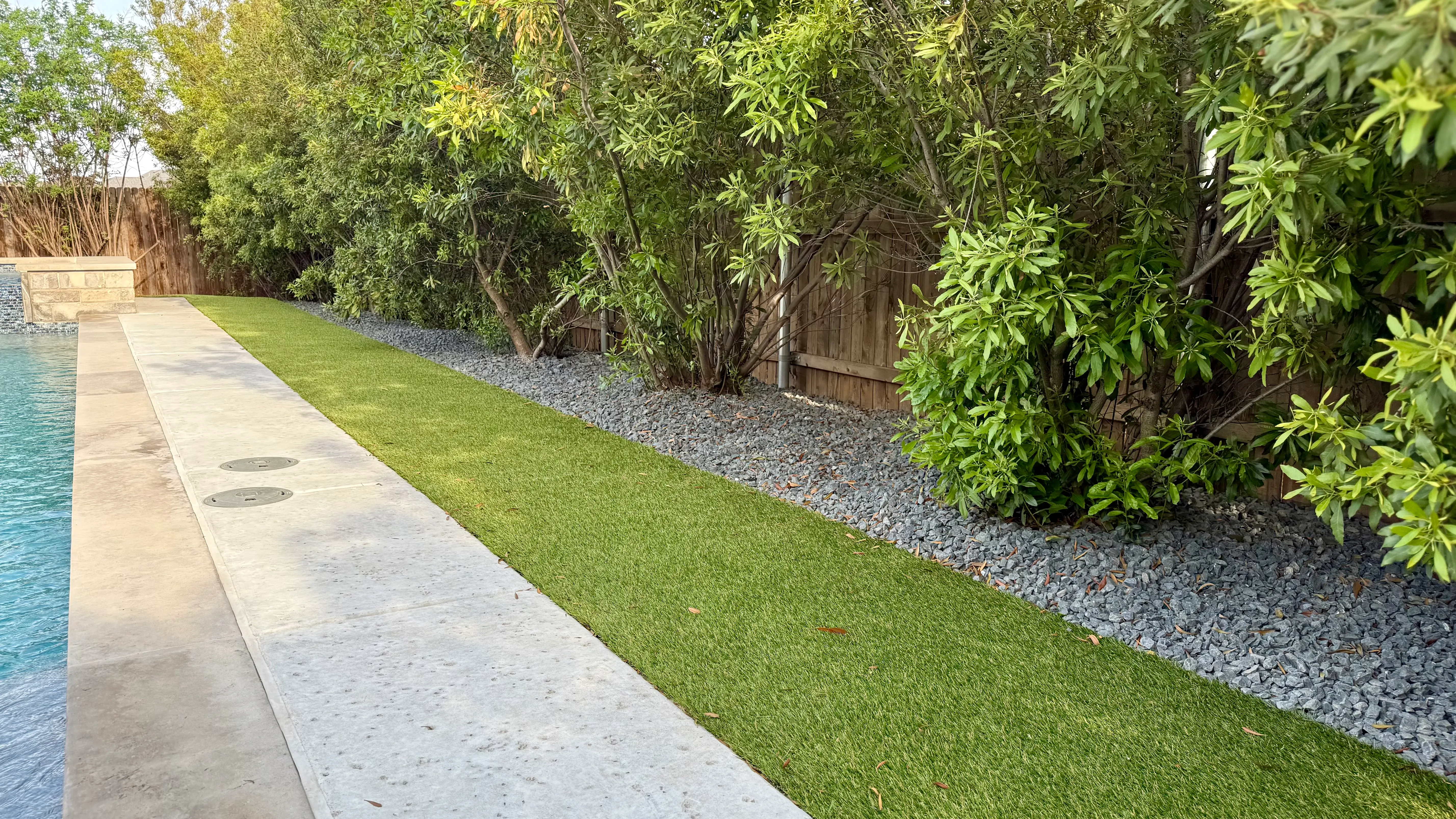 Artificial turf strip beside a pool deck bordered by a gravel landscape bed and mature bushes in DFW