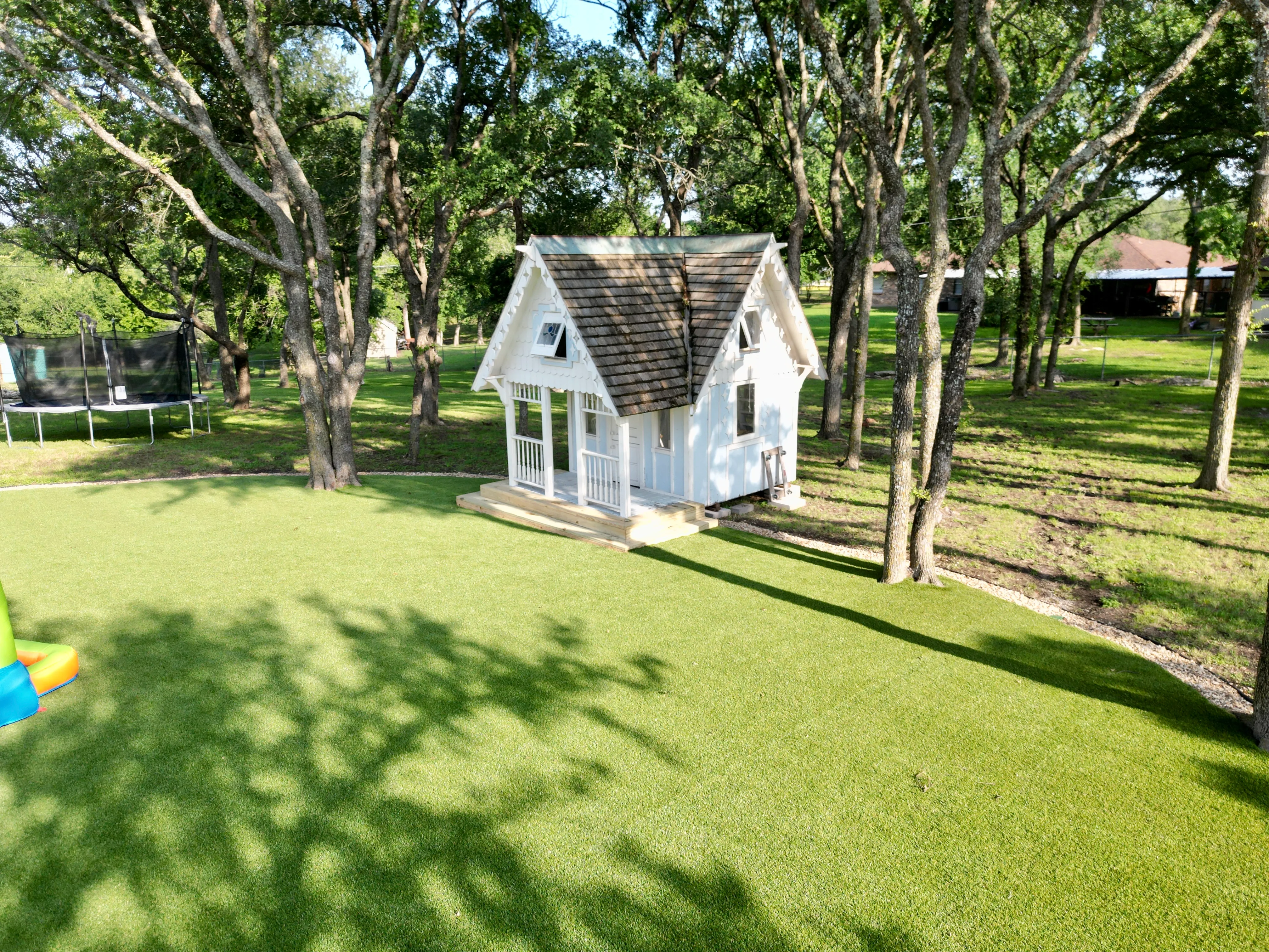 Wide view of a turf backyard with a white playhouse and trampoline nestled among mature oak trees in DFW