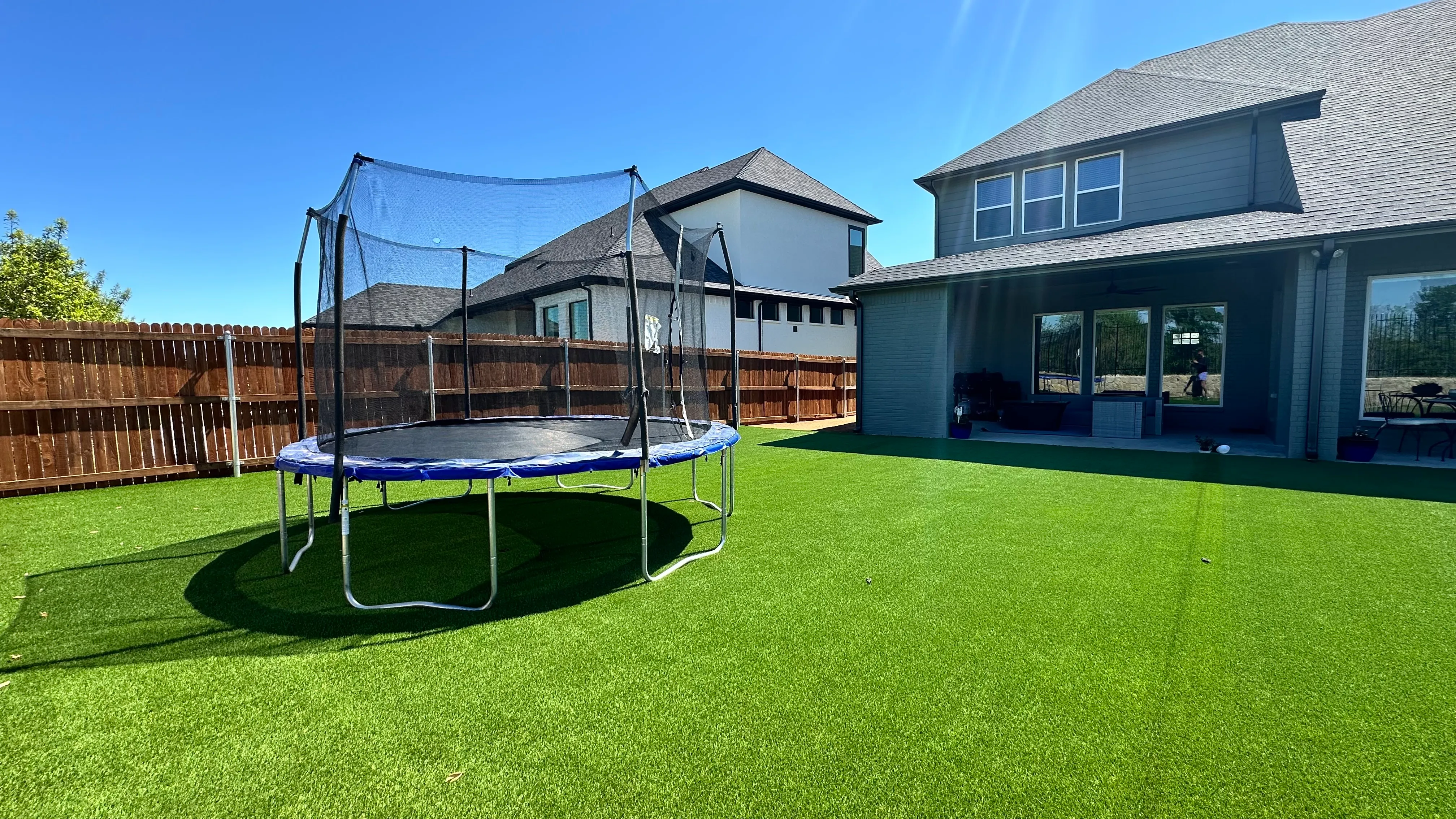 Trampoline with safety net set on an artificial turf lawn beside a gray two-story home in DFW
