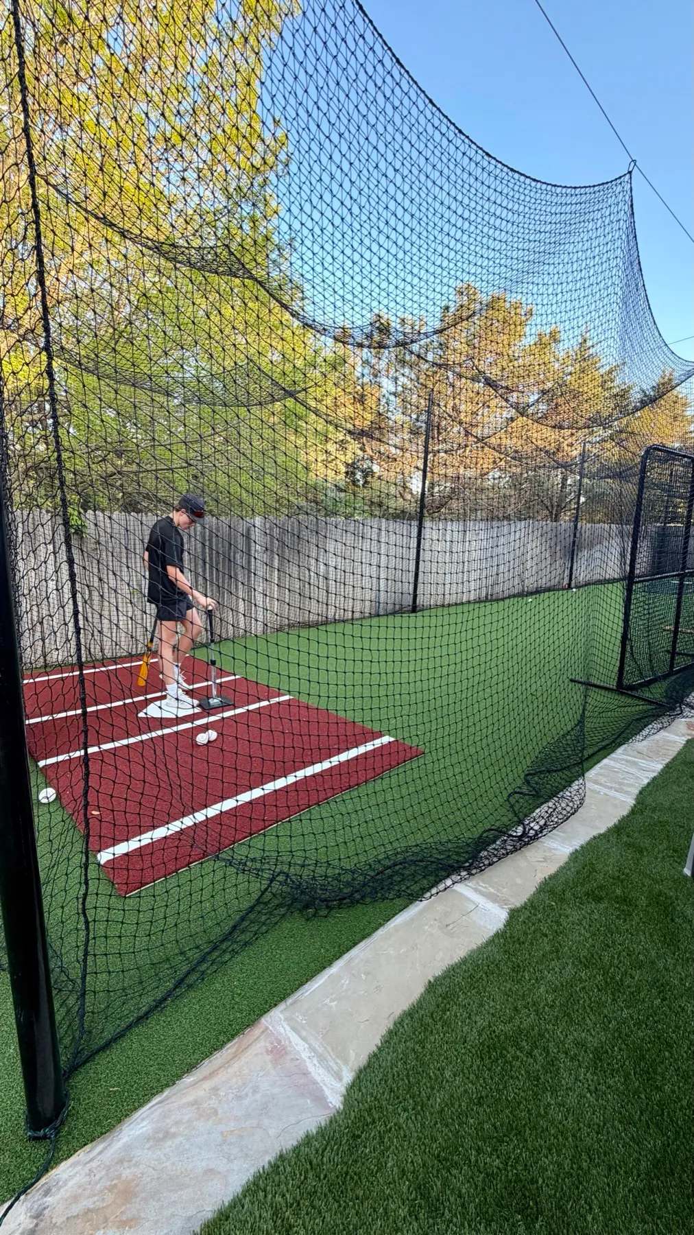 Youth batter preparing to swing inside backyard batting cage with red turf home plate mat in Aledo TX