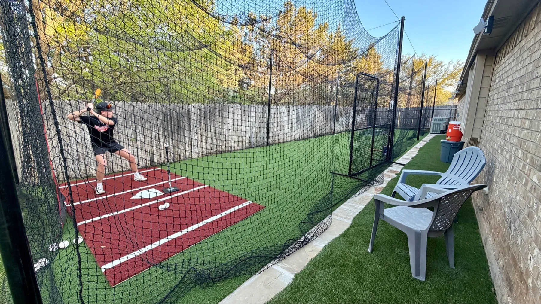 Batter mid-swing with orange bat in backyard batting cage with red turf home plate and netting in Aledo TX
