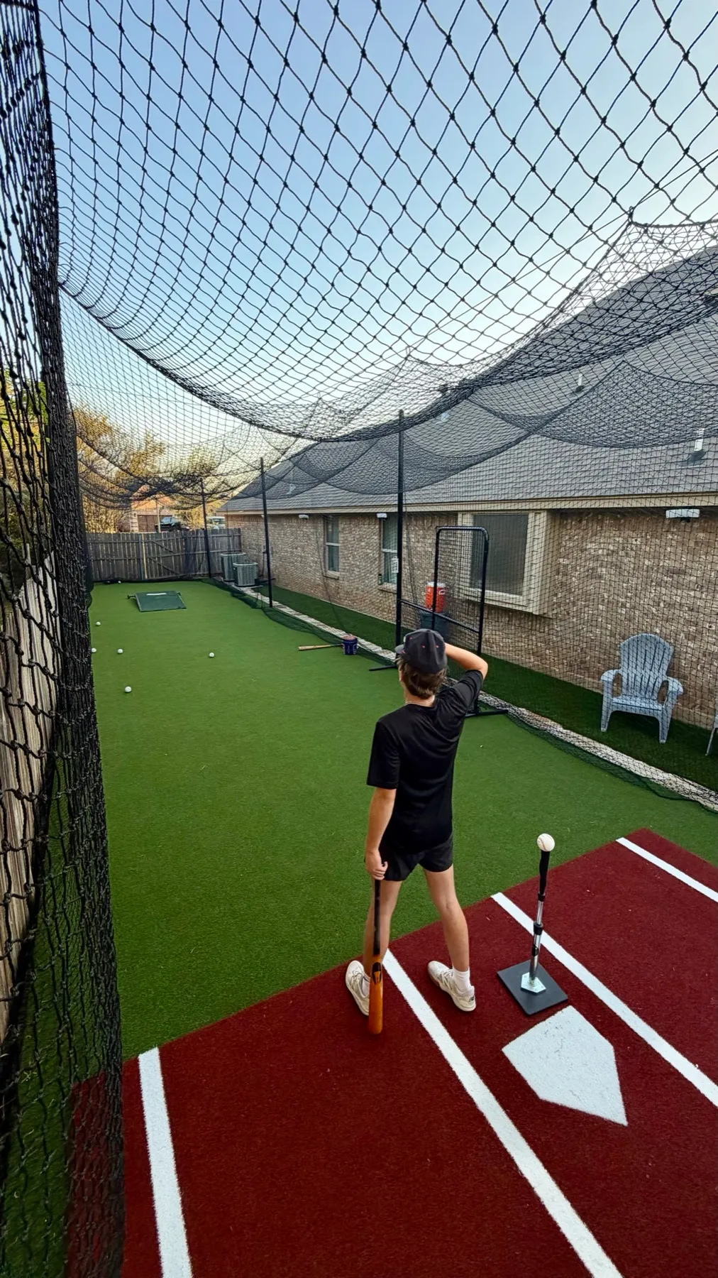 Batter with wooden bat beside tee inside backyard batting cage in Aledo TX