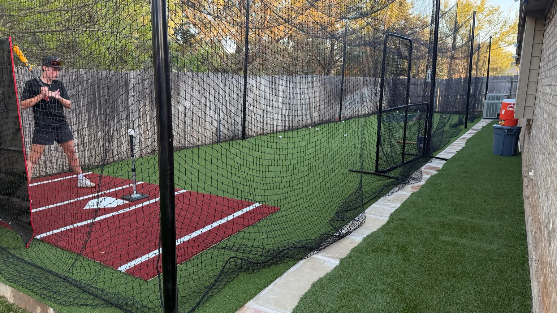 Pitcher throwing from mound end of backyard batting cage with red turf plate and flagstone walkway in Aledo TX