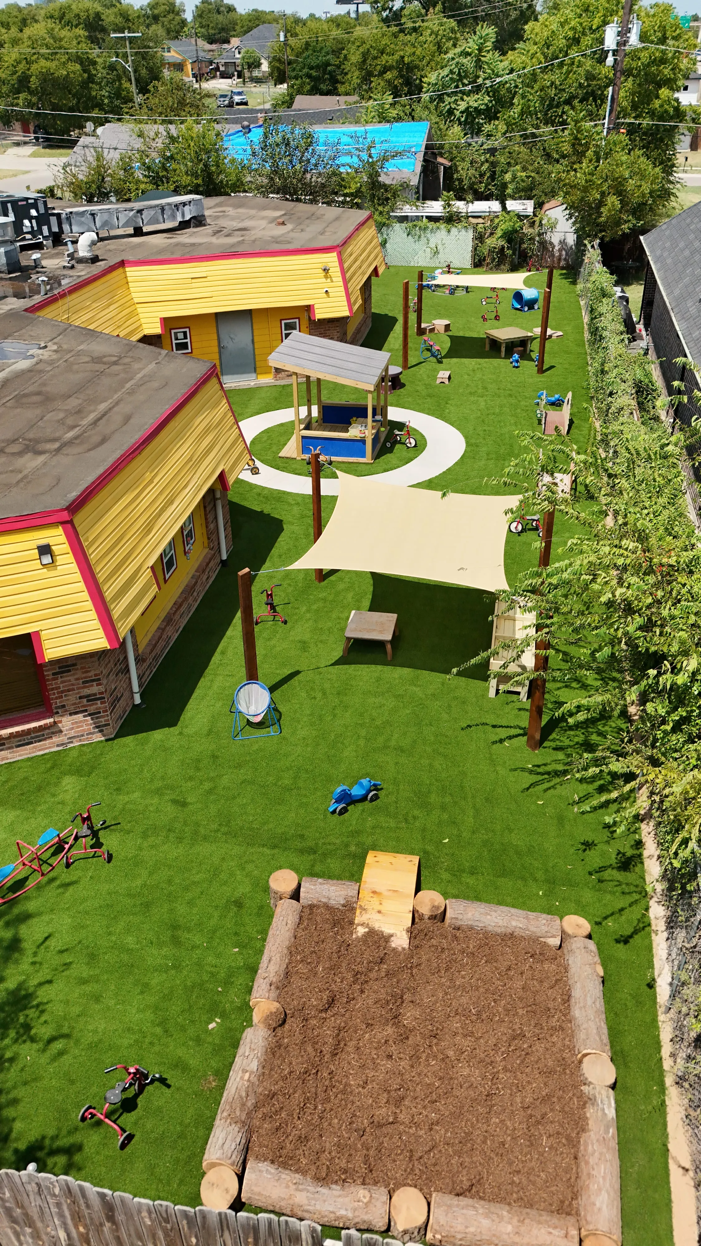 Aerial view of a daycare playground with artificial turf, shade sails, a mulch play area, and colorful ride-on toys