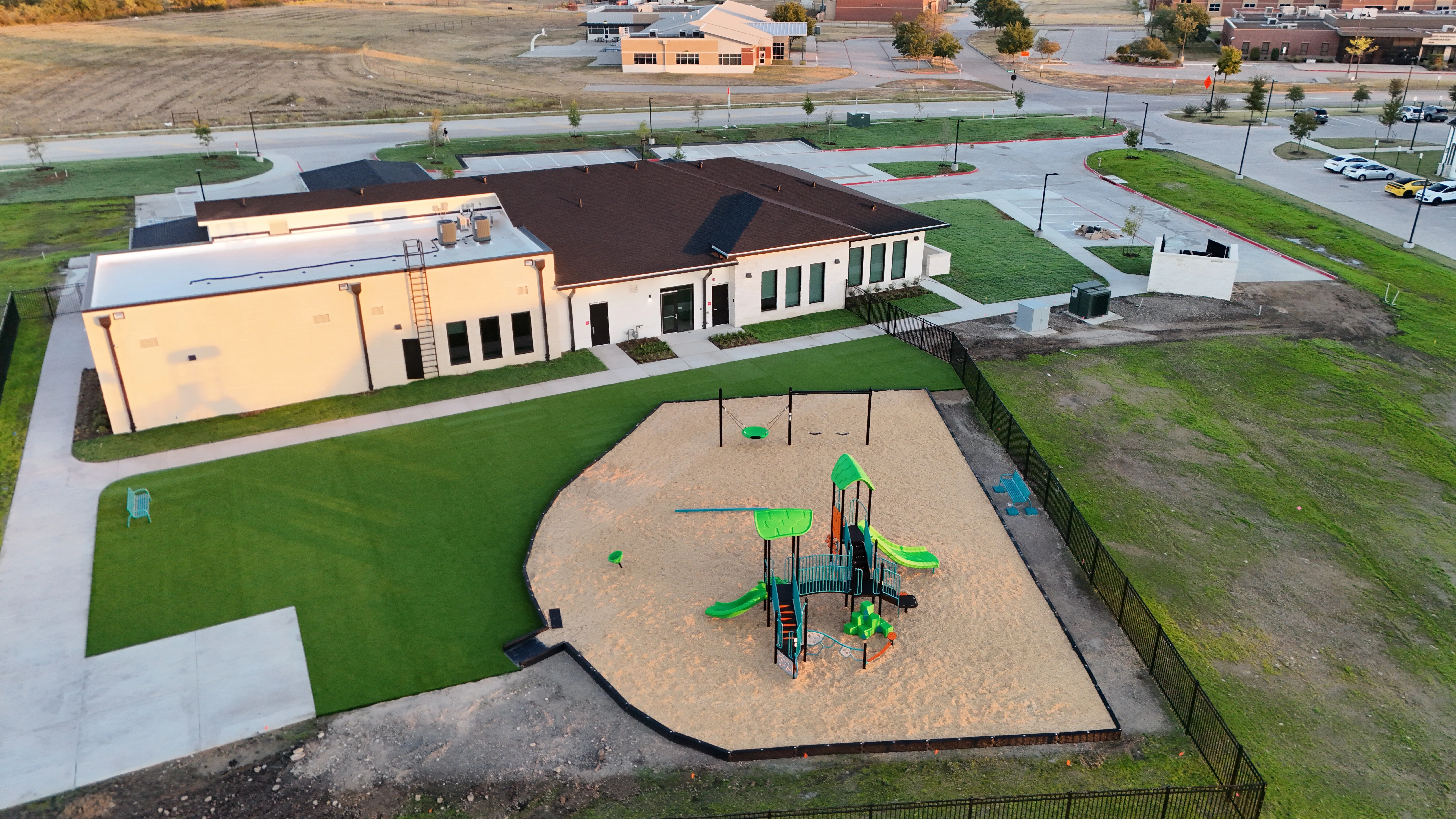 Dusk aerial of a school building with a fenced sand playground and artificial turf field
