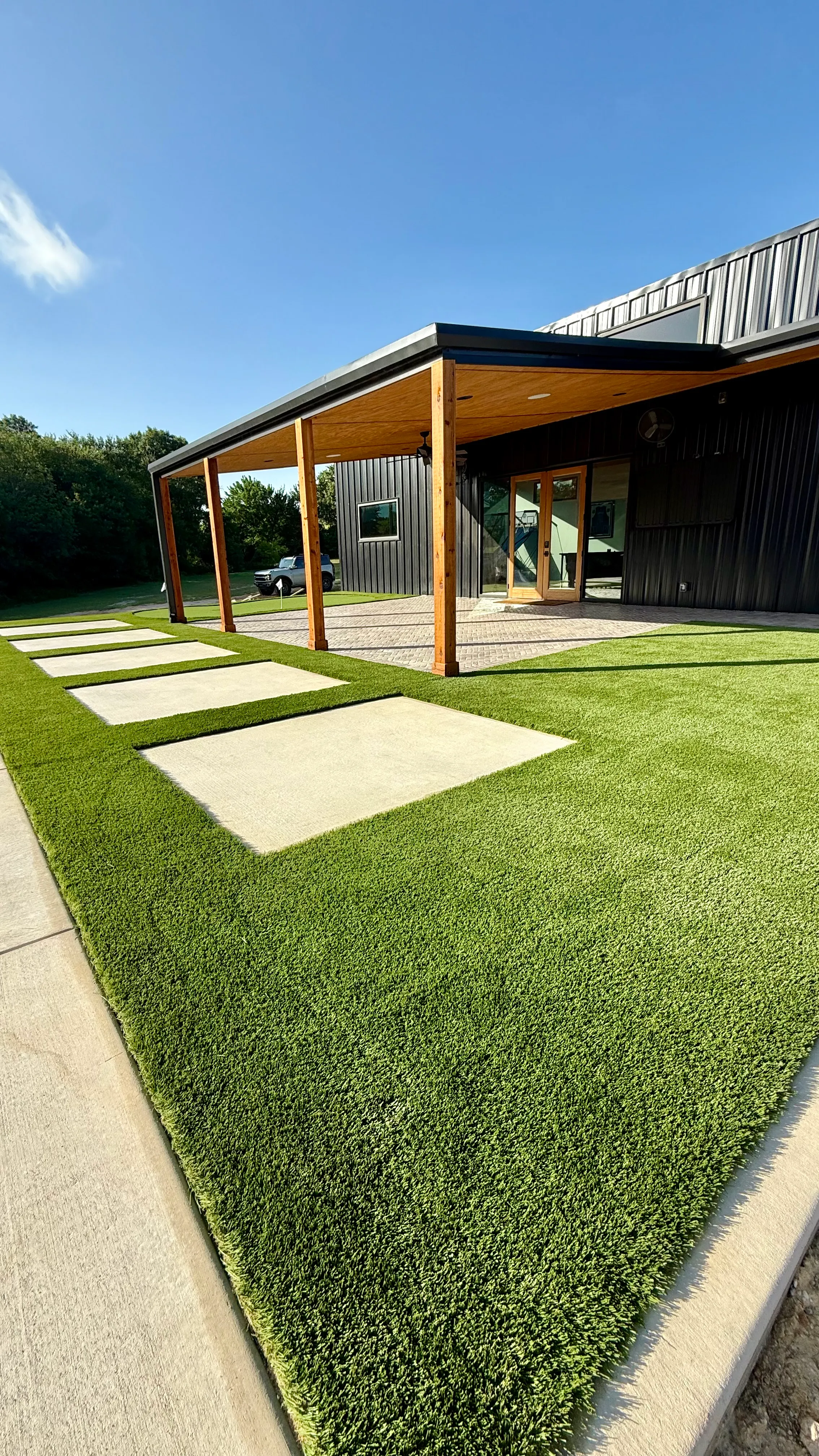 Wide view of a modern black barn-style home with turf-and-paver checkerboard pattern and a small putting green in DFW