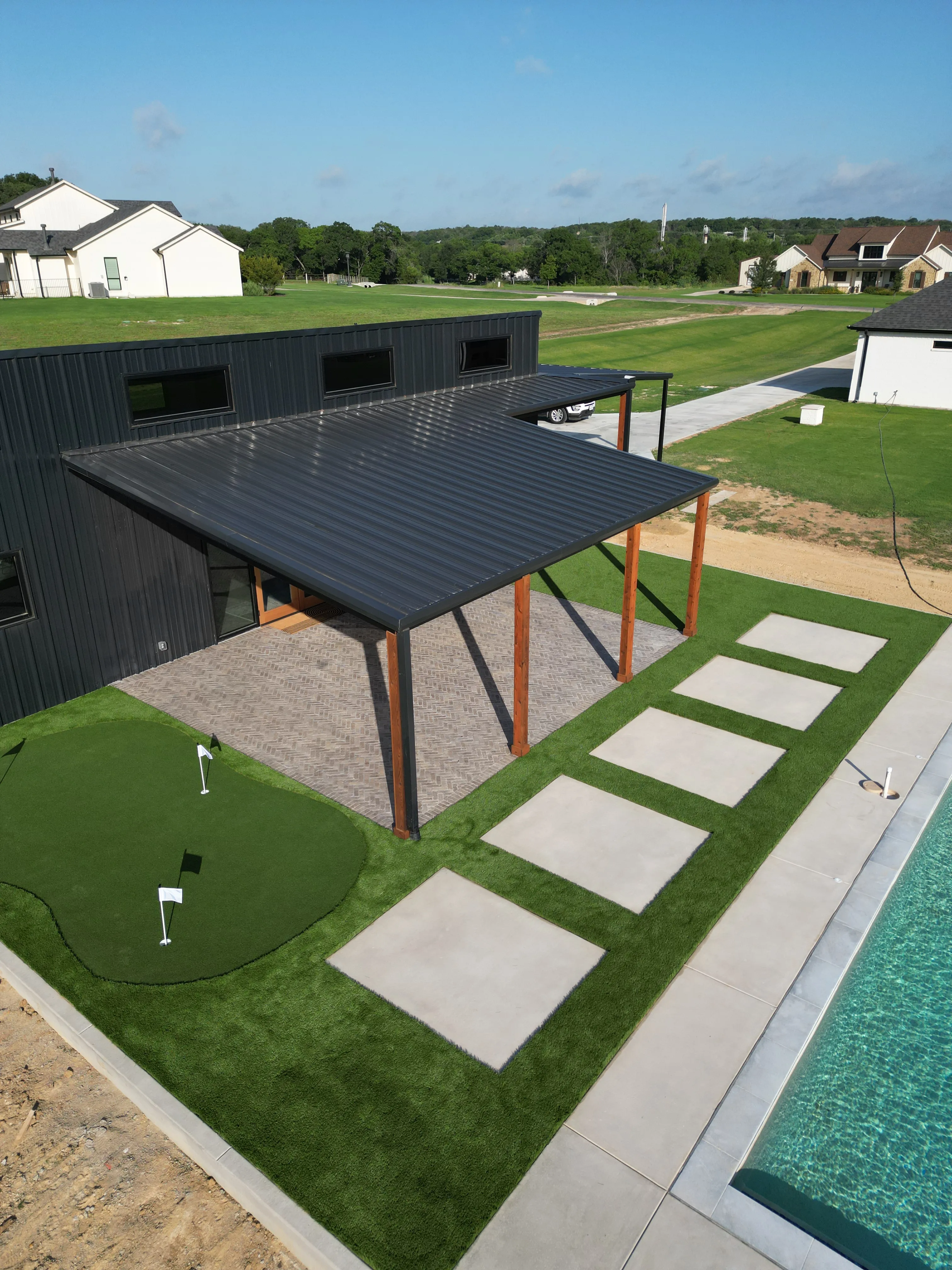 Aerial of a modern black barn-style home with a checkerboard turf and paver pad, putting green, and pool in DFW