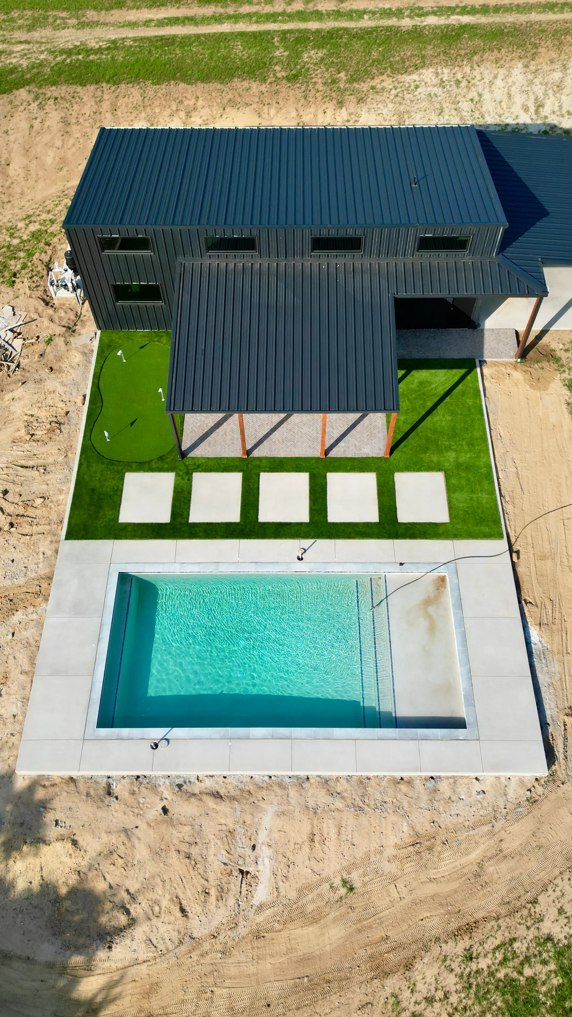Straight-down aerial of a modern home showing a checkerboard turf pad, putting green with flags, and rectangular pool in DFW