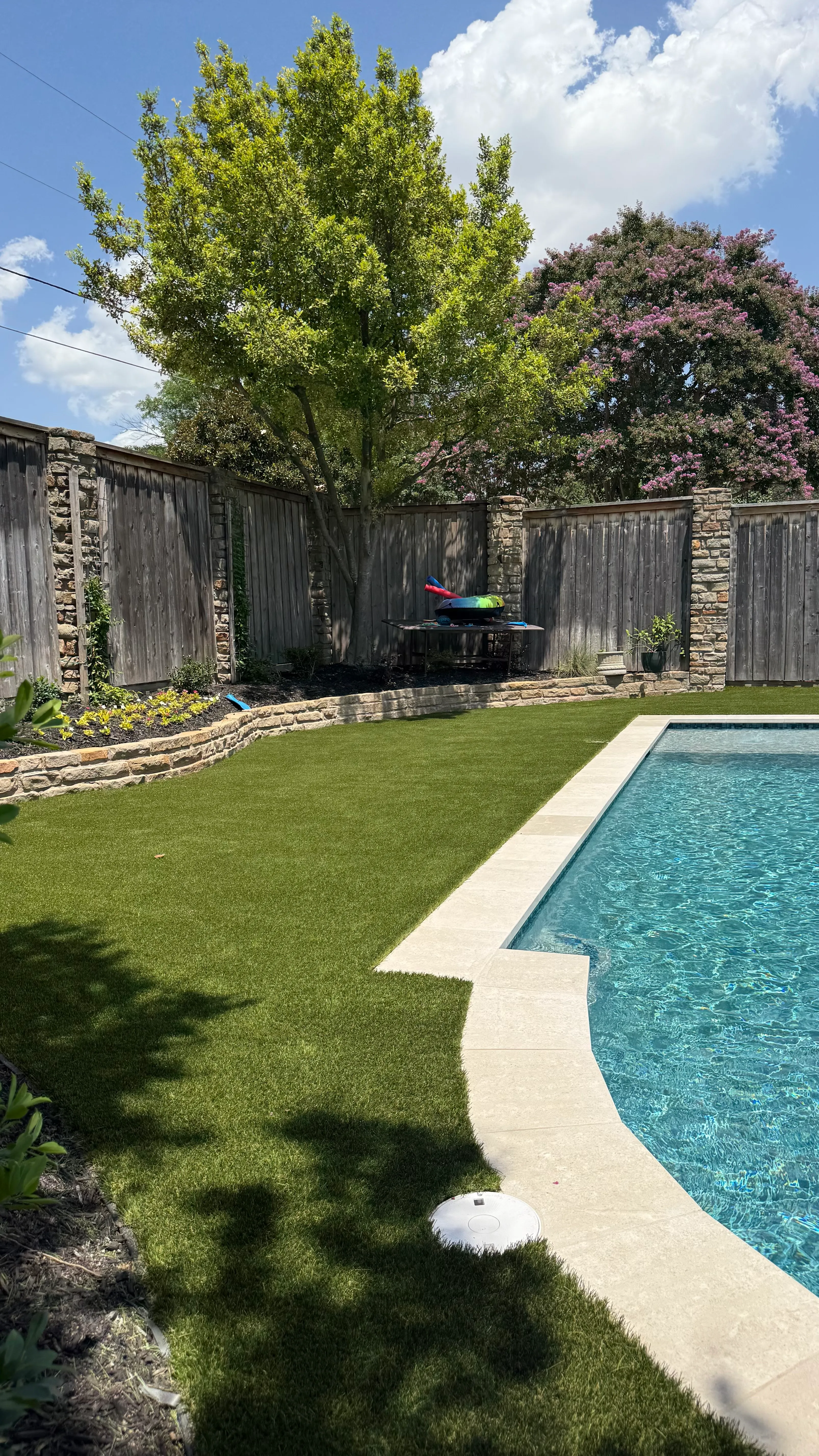 Wide view of a turf backyard with kidney-shaped pool, stone-topped wood fence, and landscaped beds in DFW