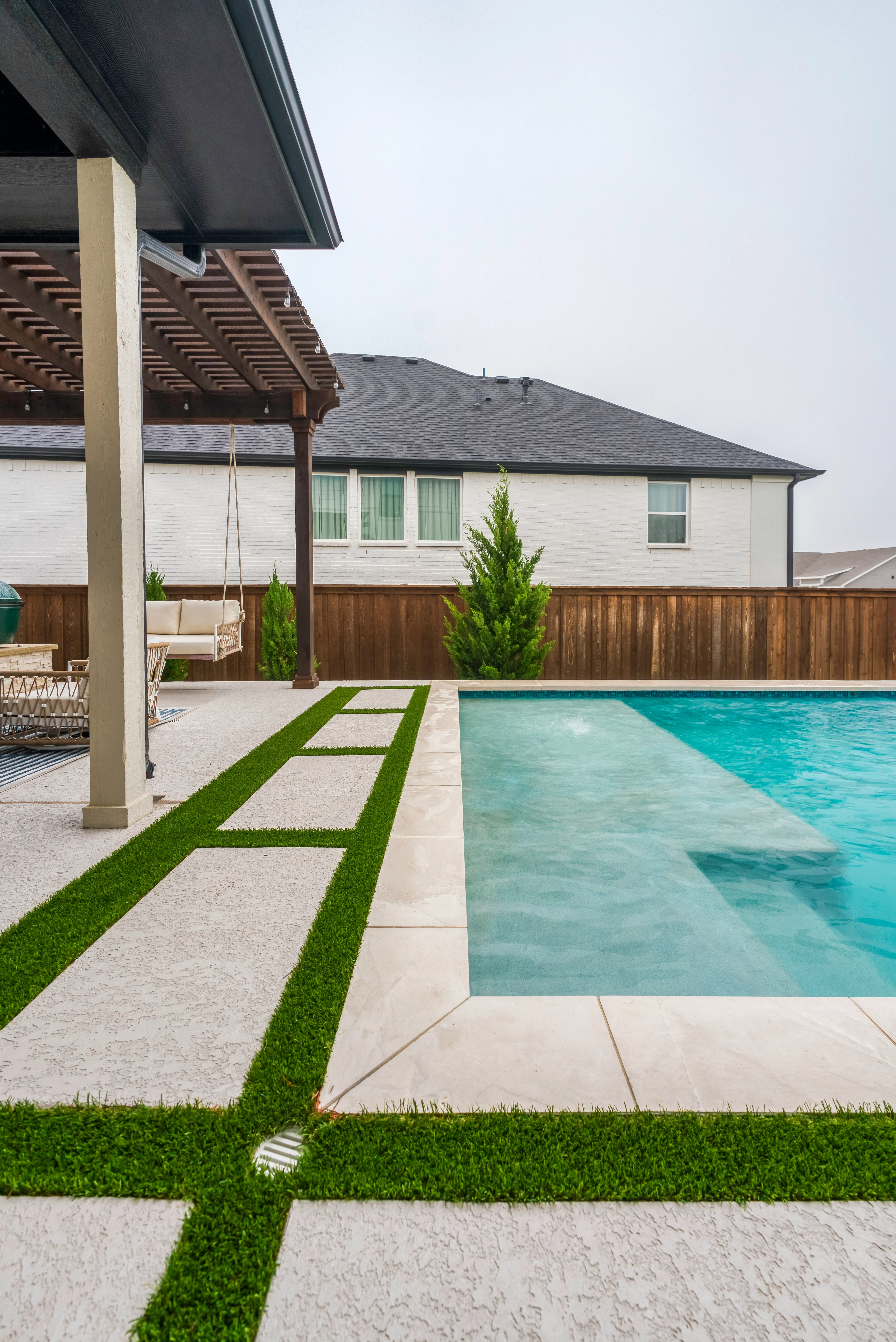 Turf strips running between large-format pavers along a pool edge, with a wood pergola and outdoor kitchen in the background
