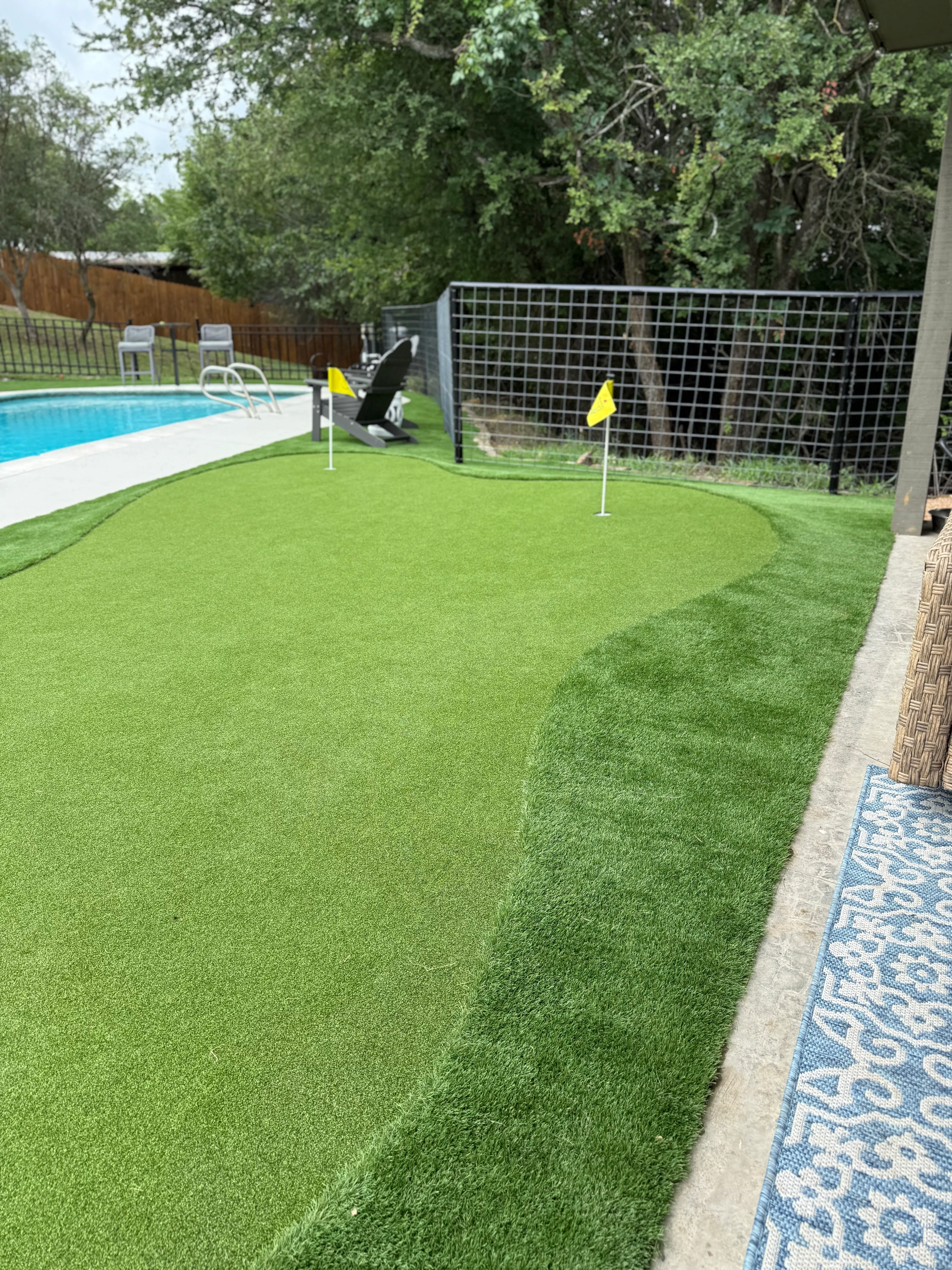 Two-hole backyard putting green with yellow flags beside a pool in DFW, fringe and green surface contrast visible