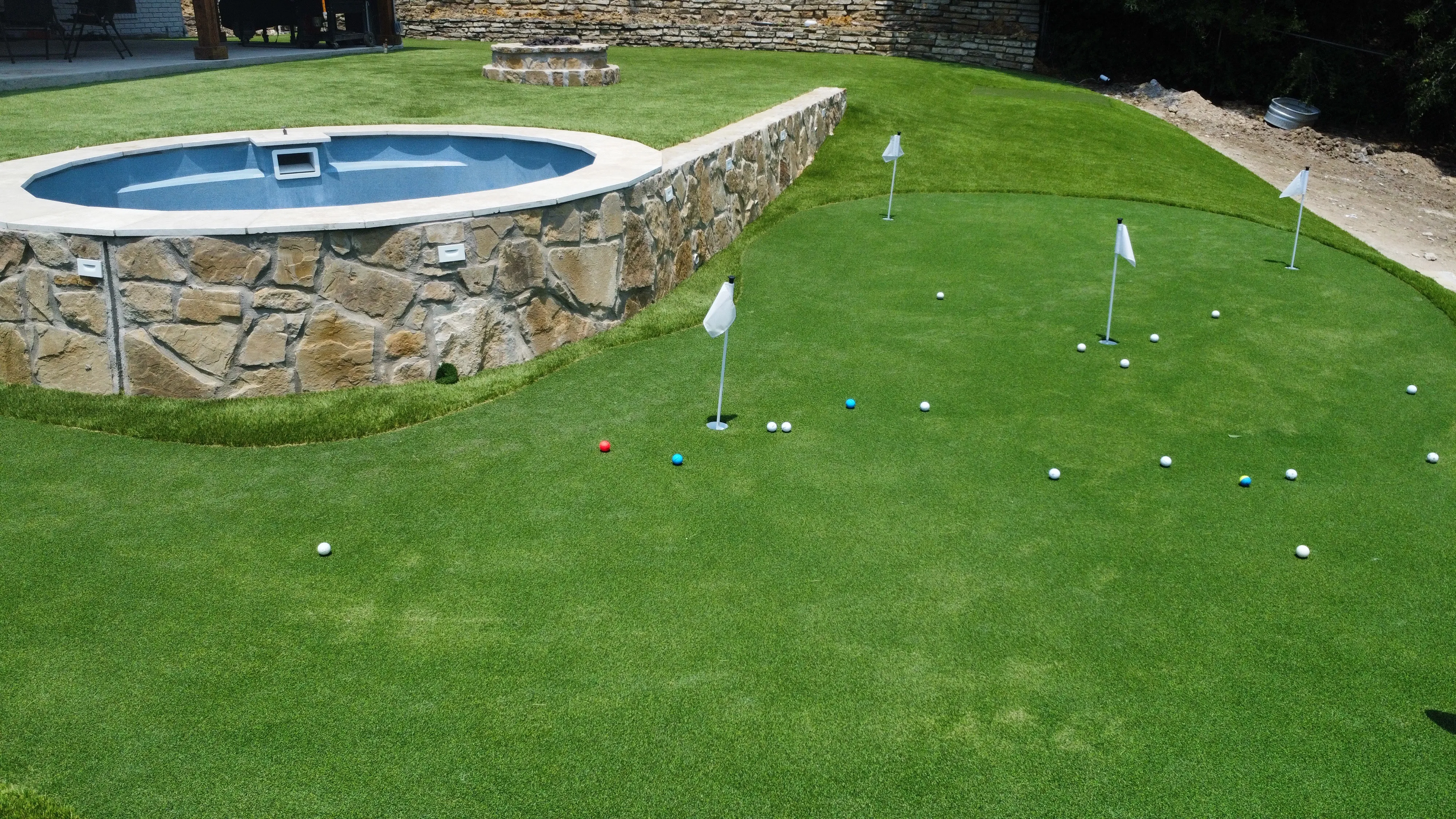 Aerial of a putting green with multiple flags and golf balls beside a raised stone-walled pool in DFW