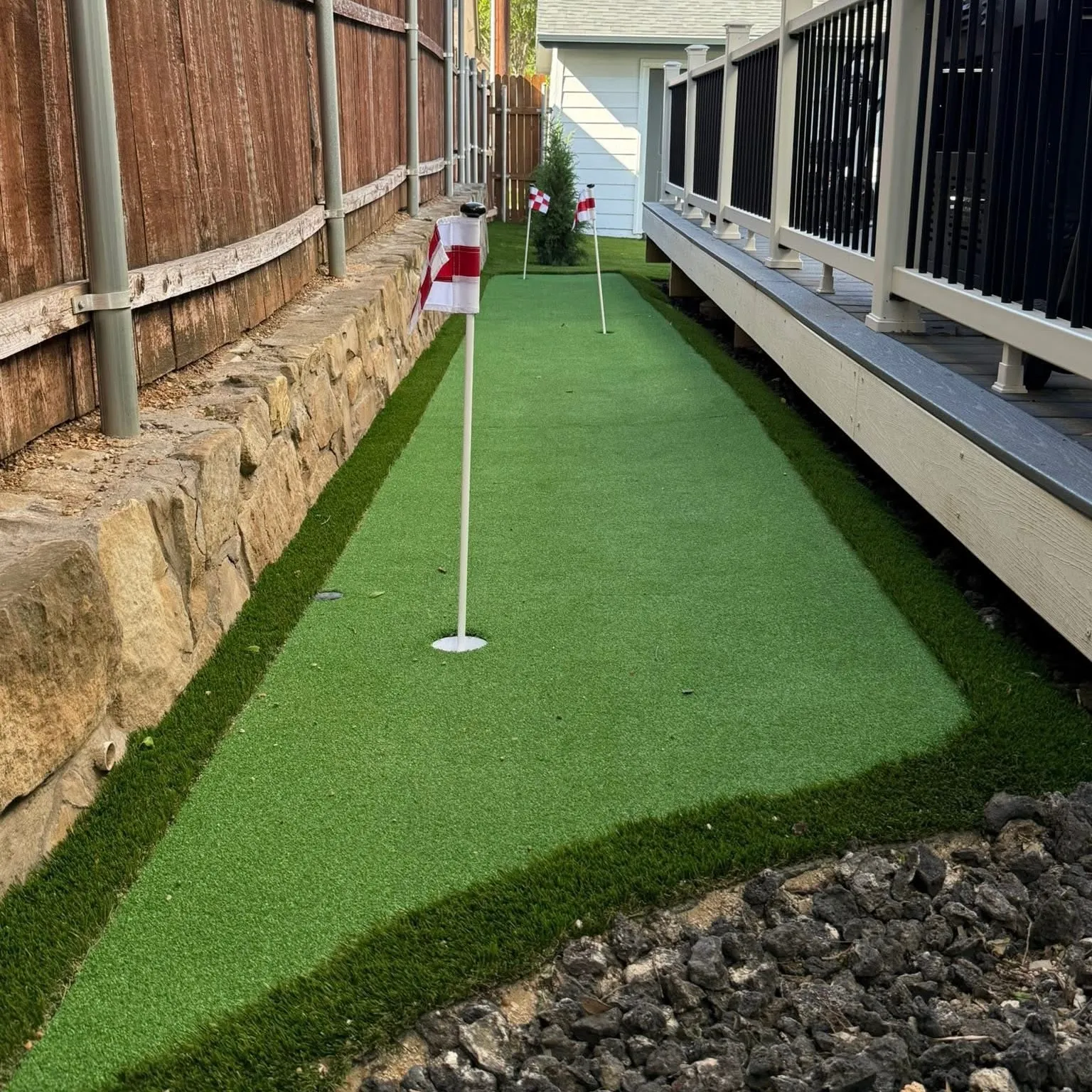 Narrow backyard putting green with two flags beside a stone retaining wall and deck in DFW