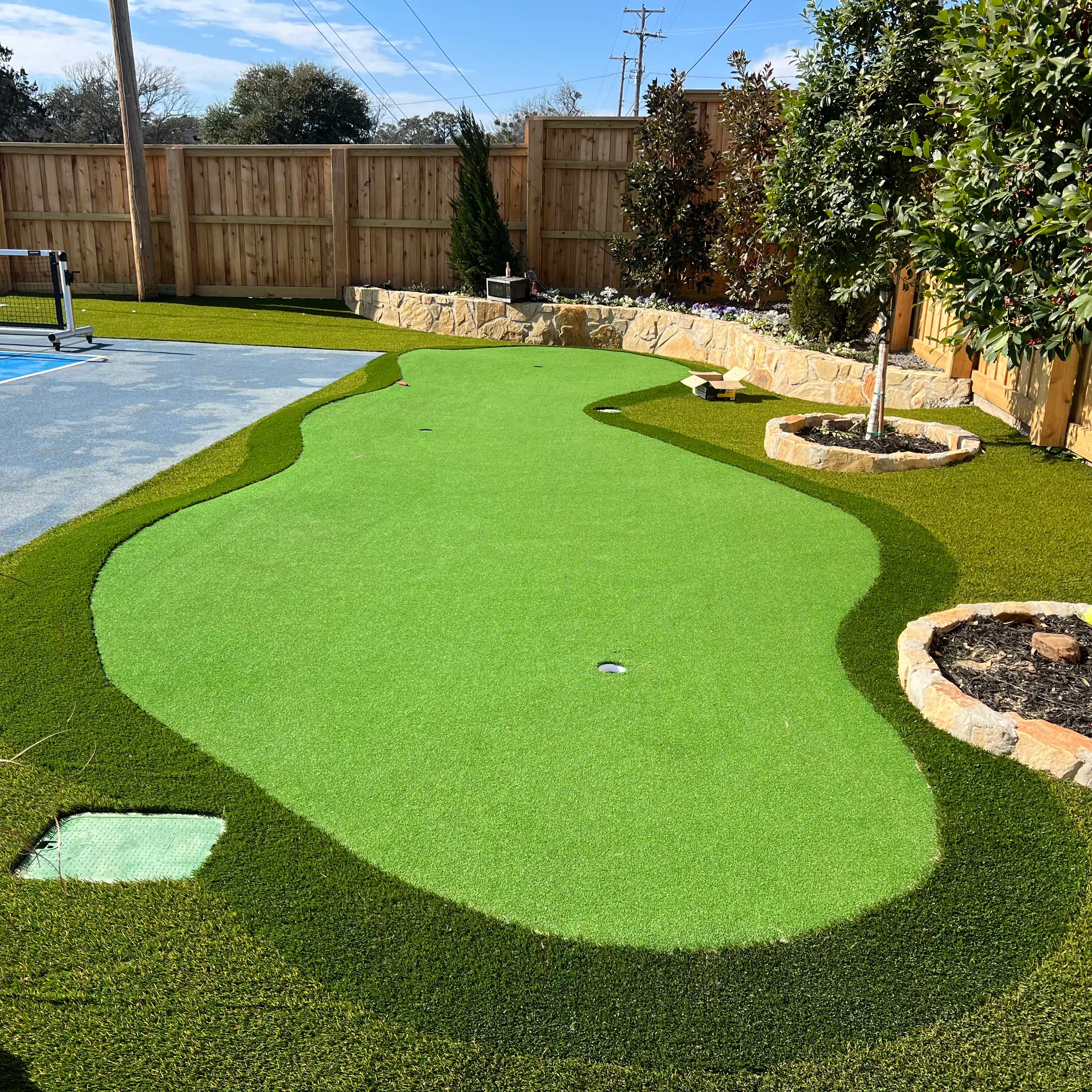 Curved amoeba-shaped backyard putting green with multiple holes, stone tree rings, and a fire pit area in DFW
