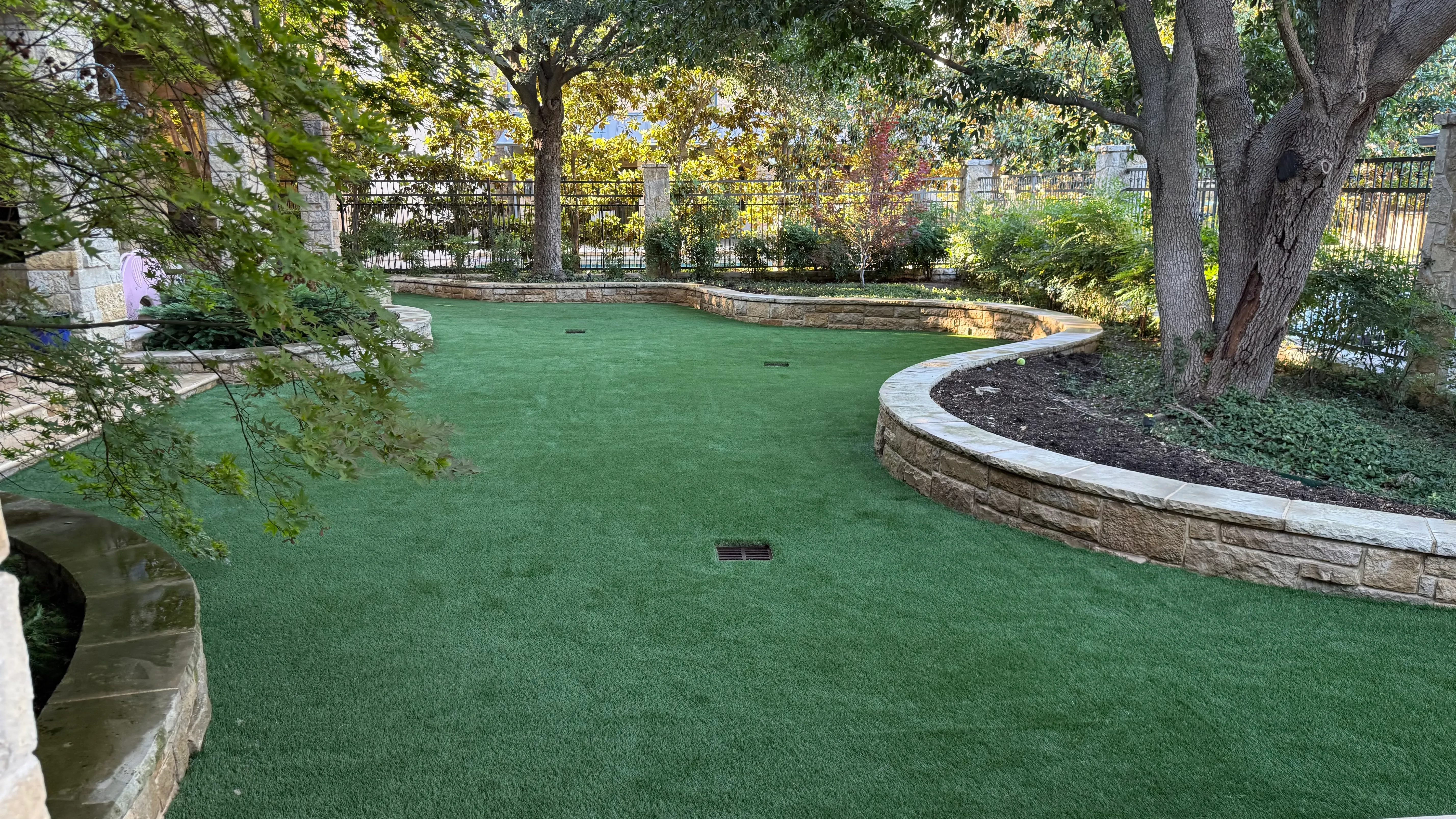 Backyard putting green viewed from beneath a mature oak tree toward a stone estate home in DFW