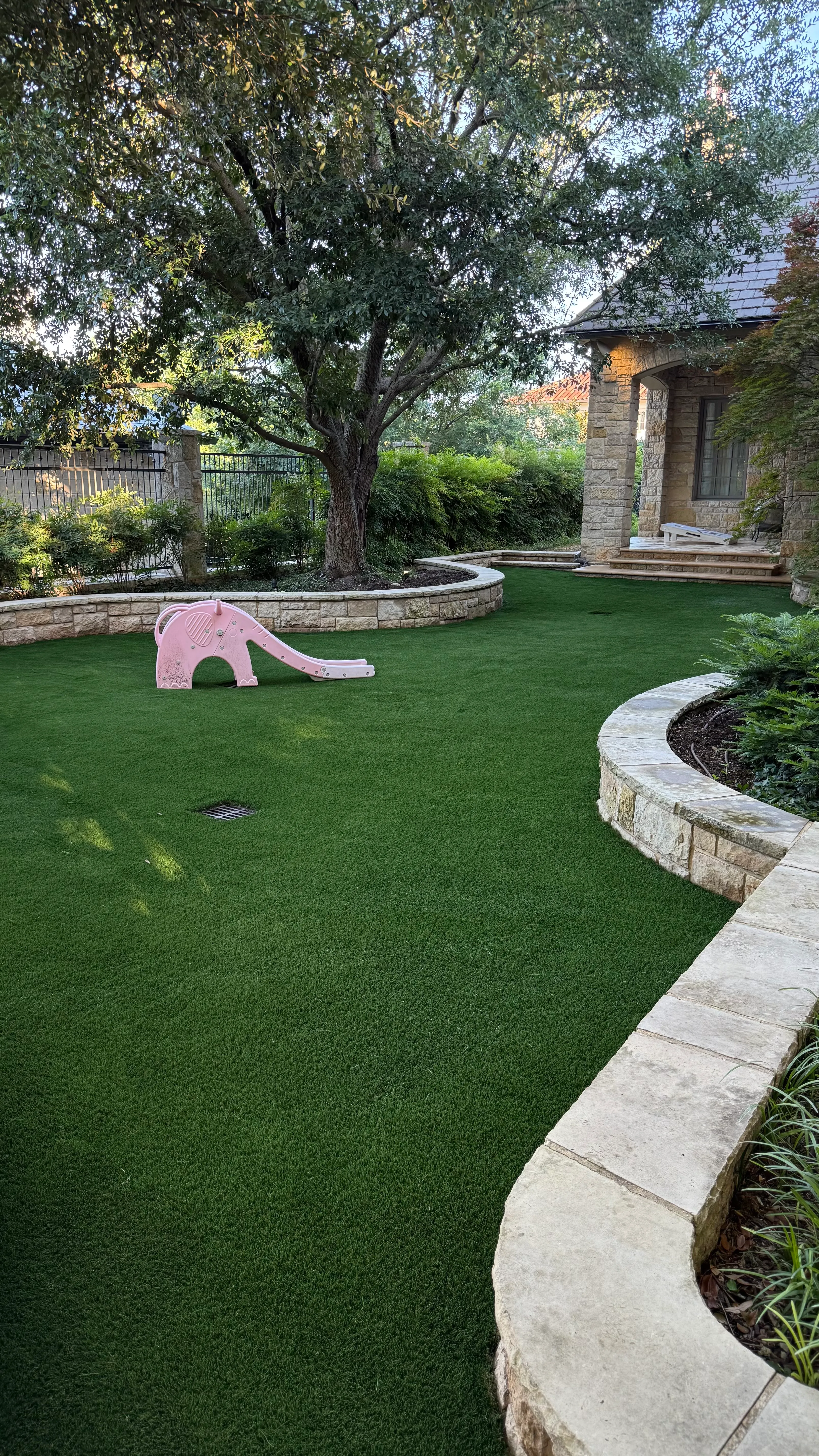 Pink toy slide sitting on a curved backyard putting green beside a stone retaining wall in DFW