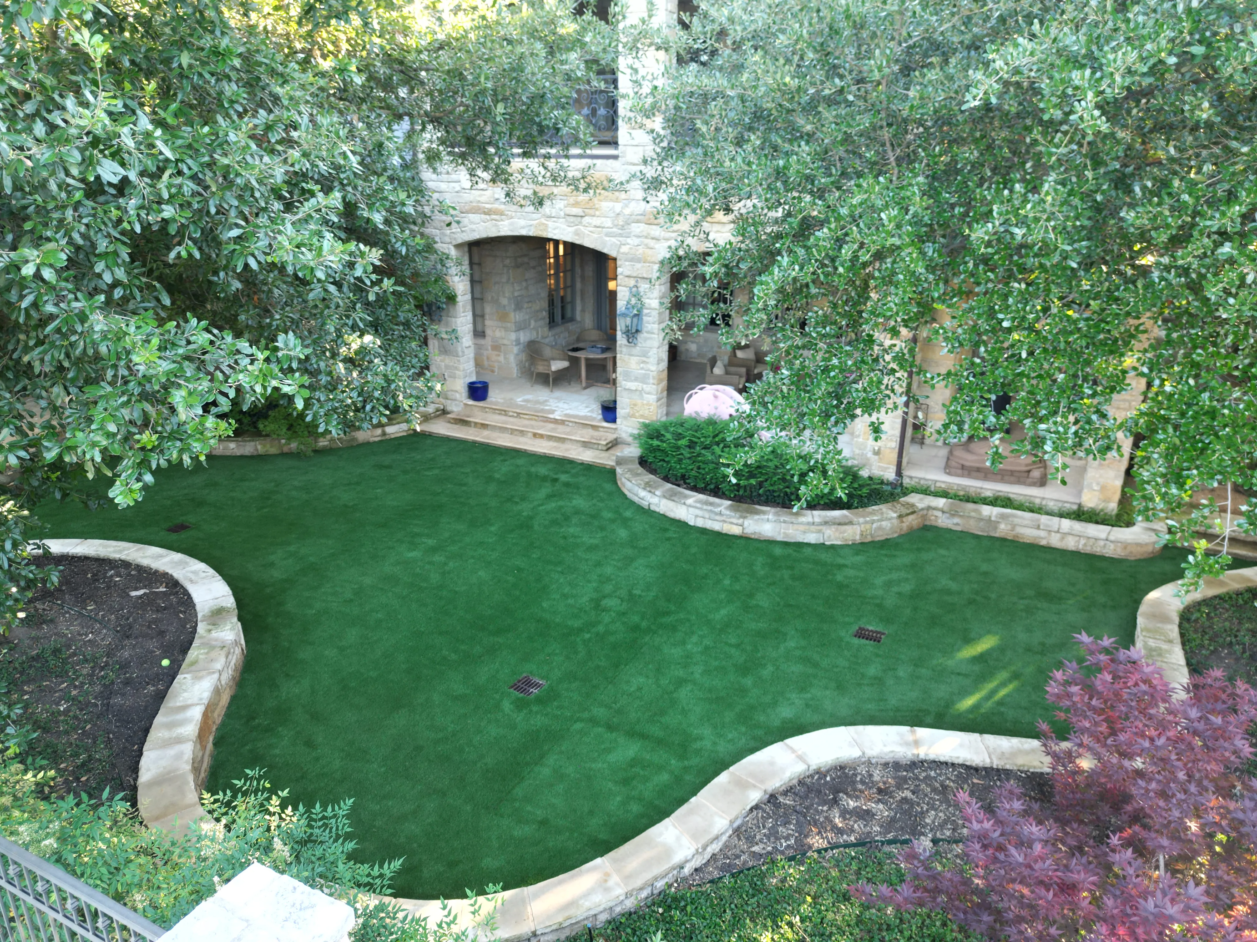 Aerial view of a curved artificial putting green wrapping around a stone courtyard and mature oak trees at a DFW estate