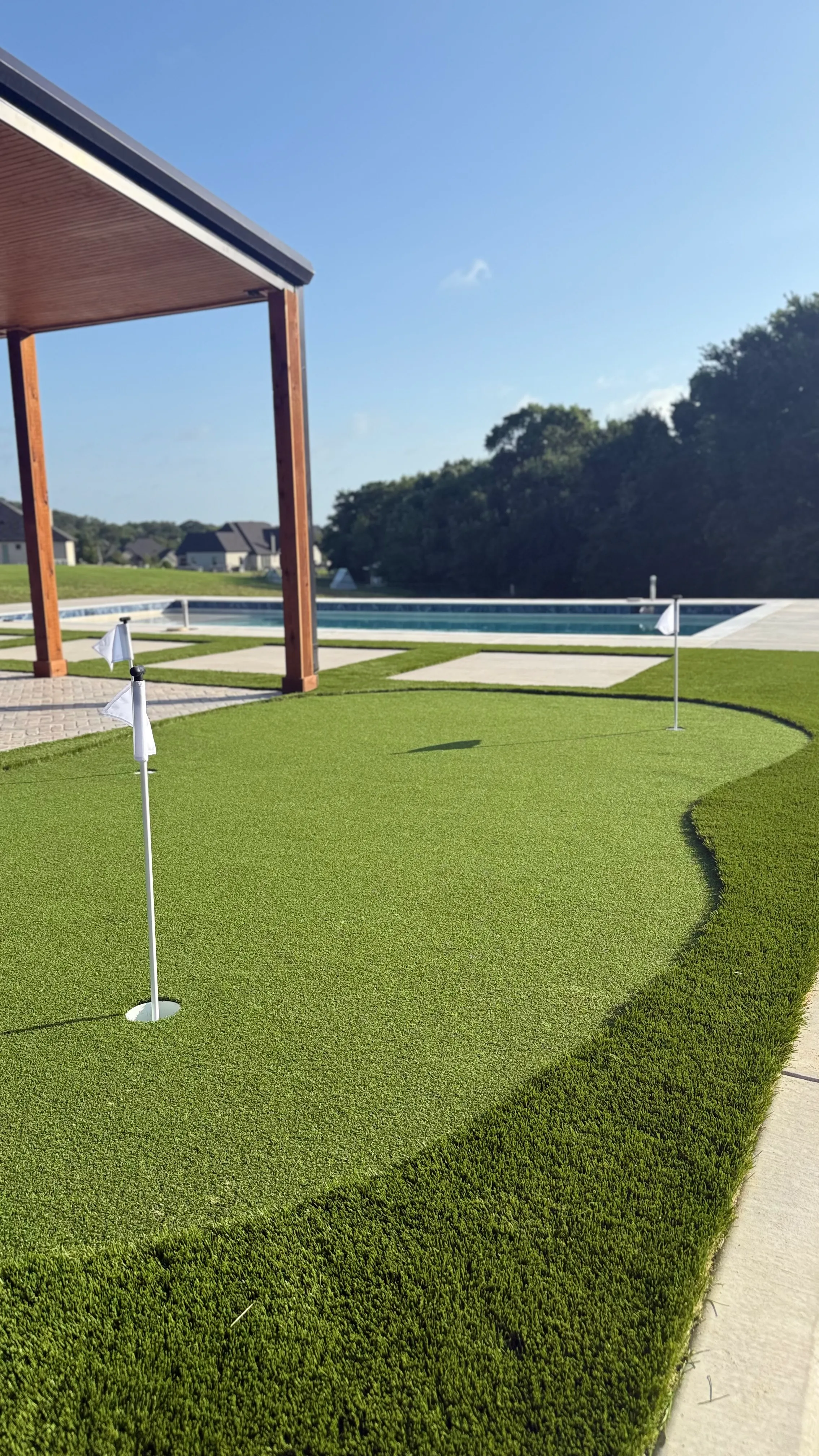 Backyard putting green with two flags beside a pool and wood pergola in a DFW yard bordered by trees