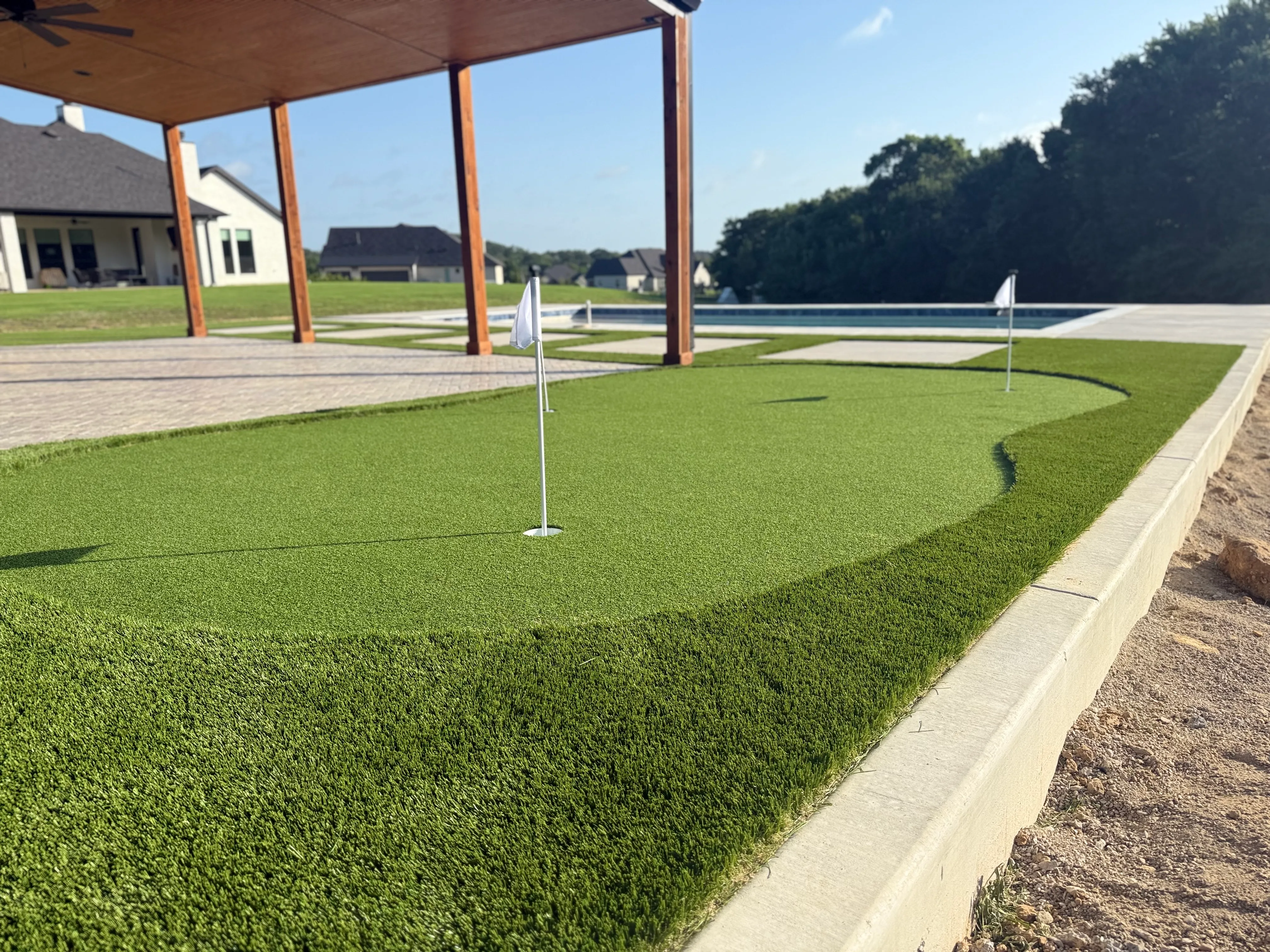 Low angle of a backyard putting green flag and hole beside a pool and covered pergola patio in DFW