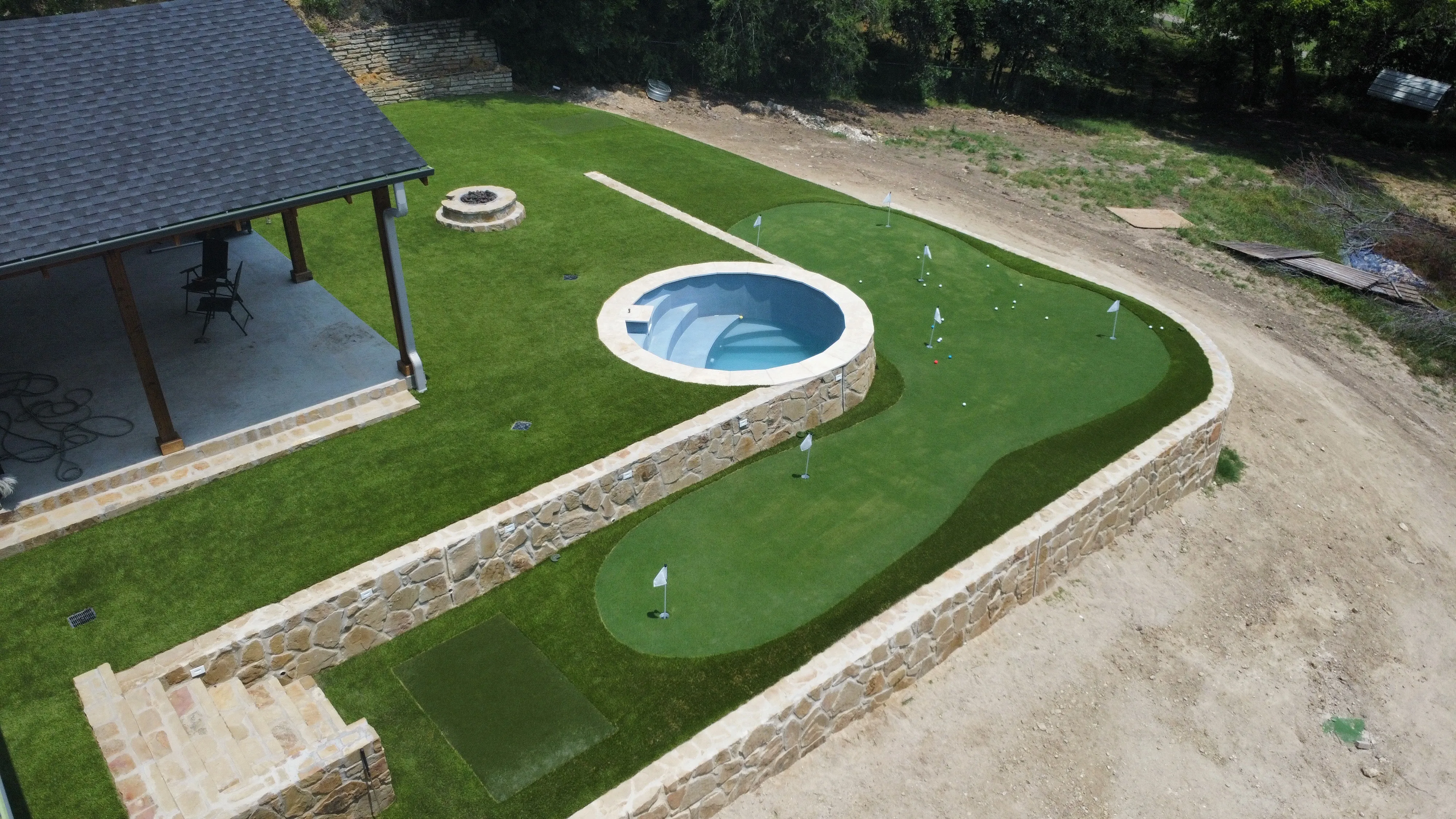 Aerial view of a large multi-flag artificial putting green beside a round pool and wood pergola patio at a DFW hillside estate
