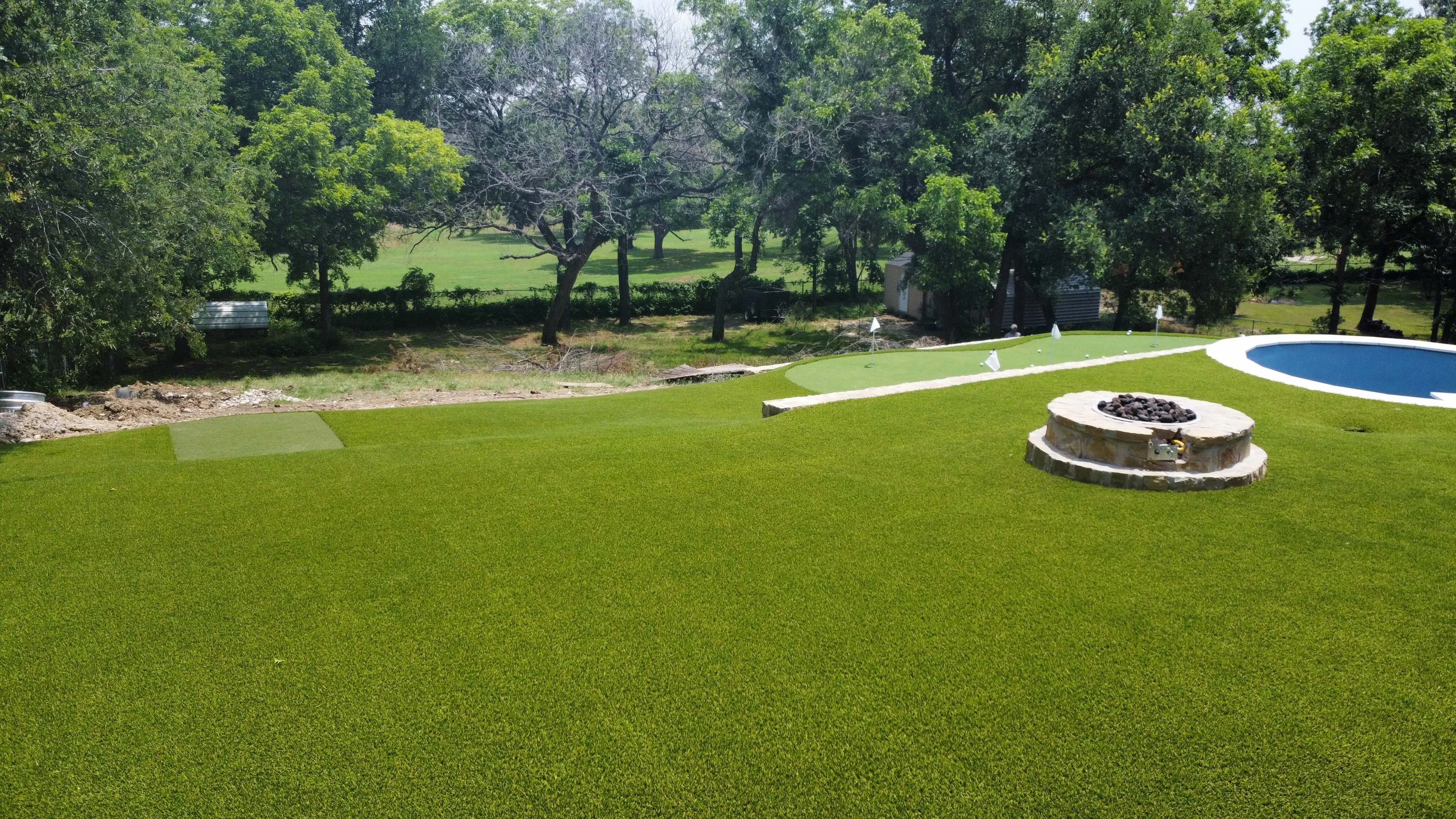 Wide view of a backyard putting green and round pool bordered by stone walls, with an open pasture visible through the tree line beyond