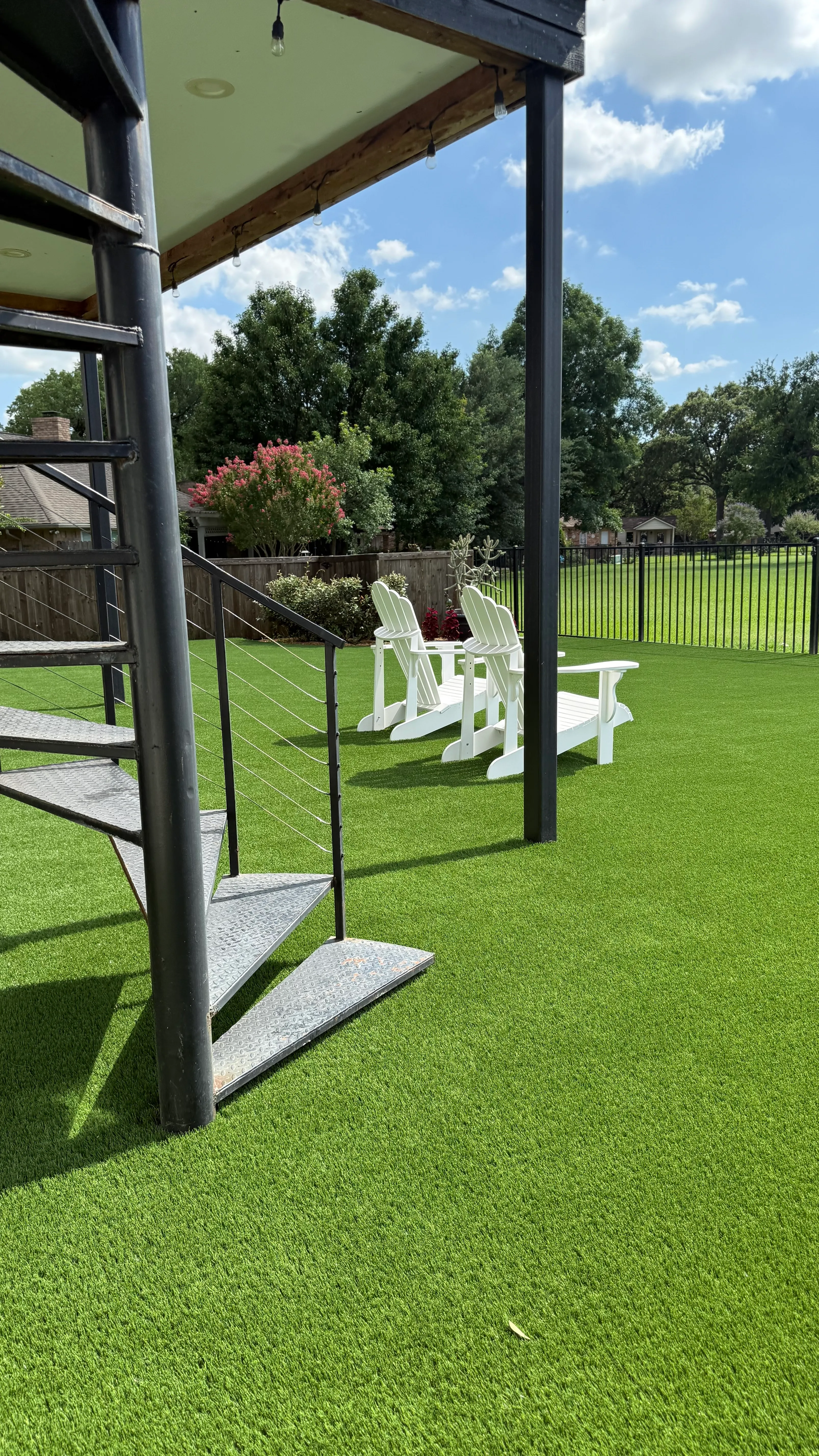 White Adirondack chairs on a turf lawn beneath a black spiral staircase leading to a rooftop deck at a DFW home