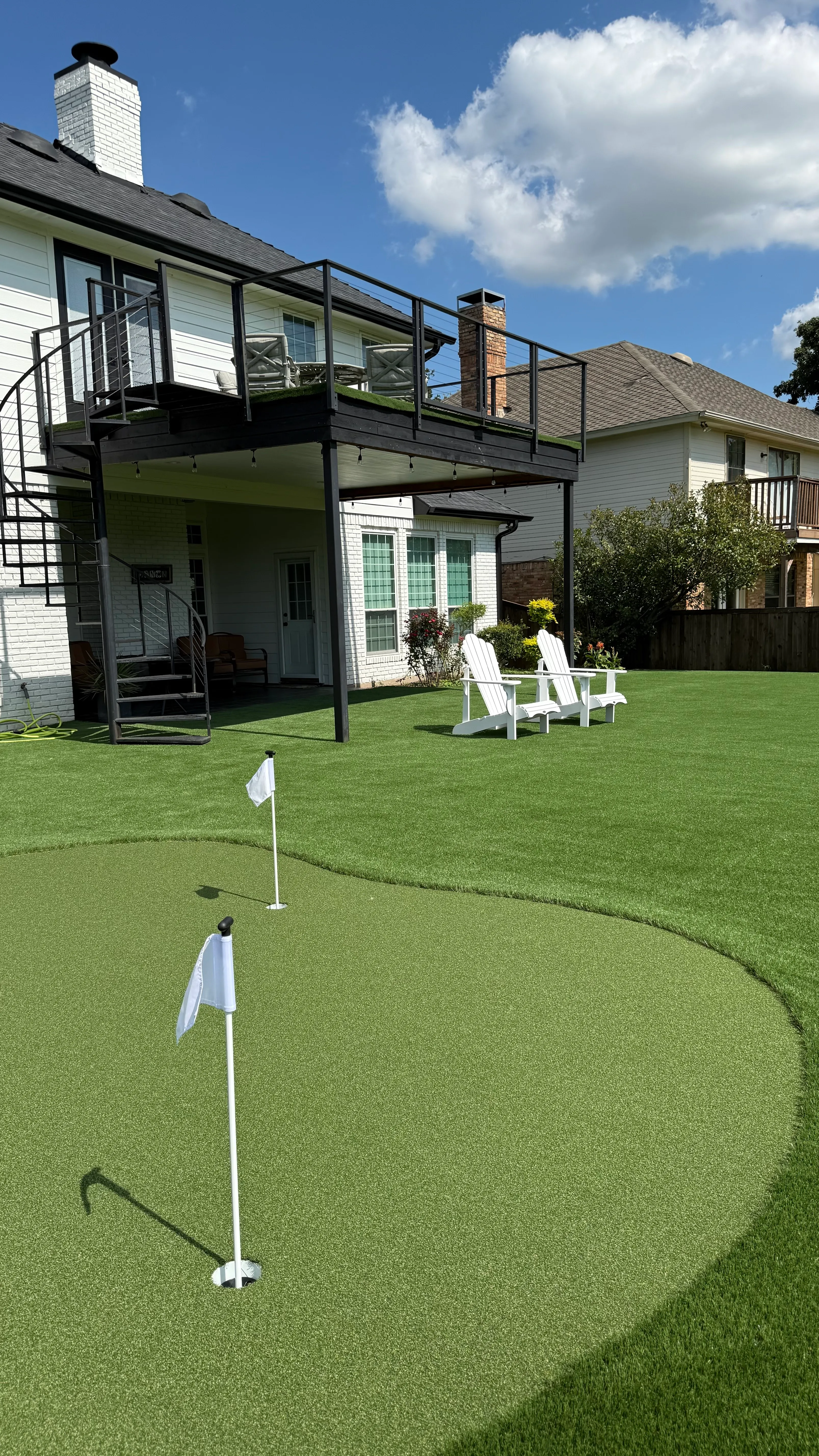 Putting green with two flags in front of a two-story home with a black spiral staircase and elevated deck in DFW