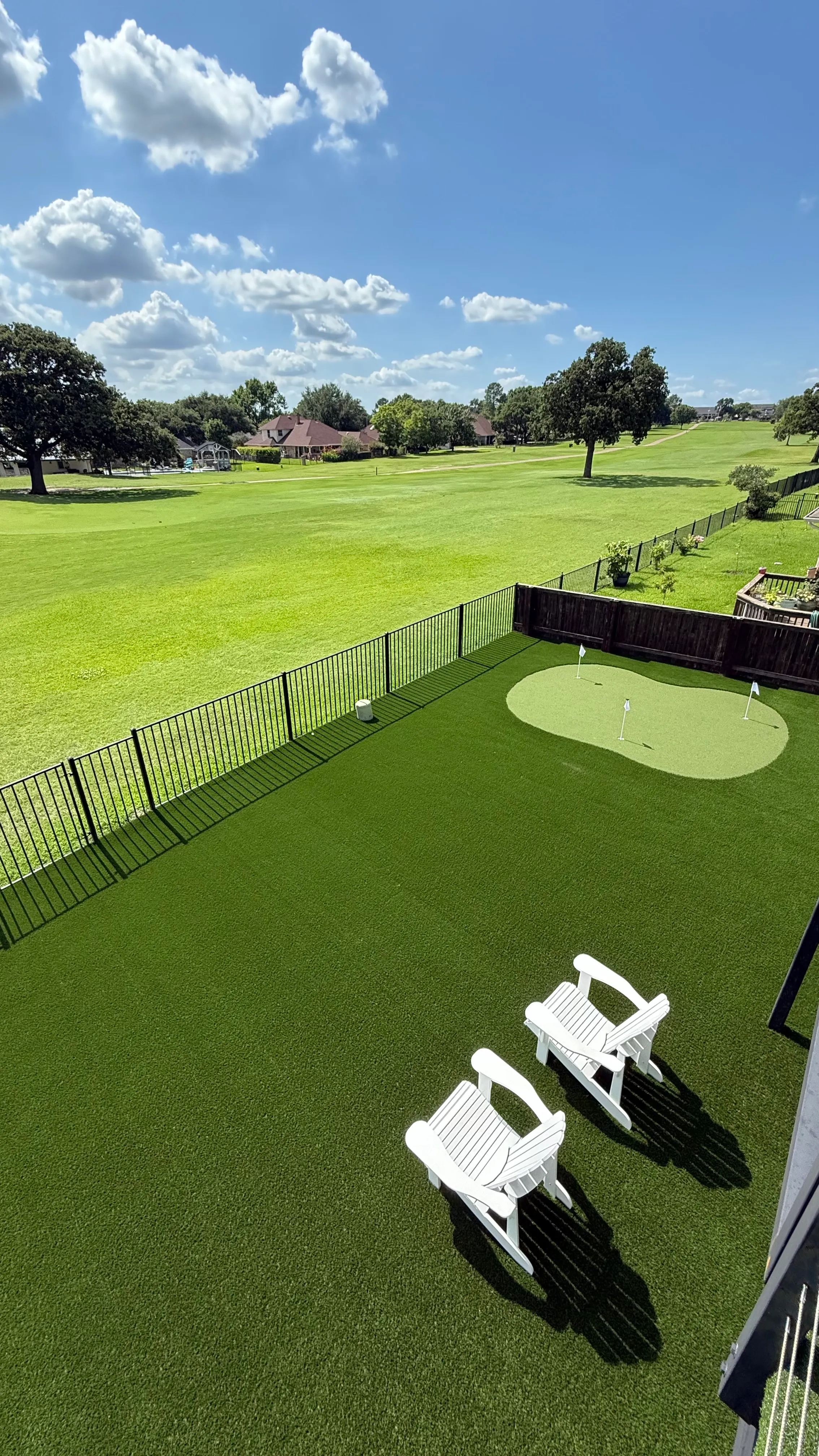 Elevated view from a rooftop deck over a putting green and iron fence toward an open lawn and distant houses in DFW