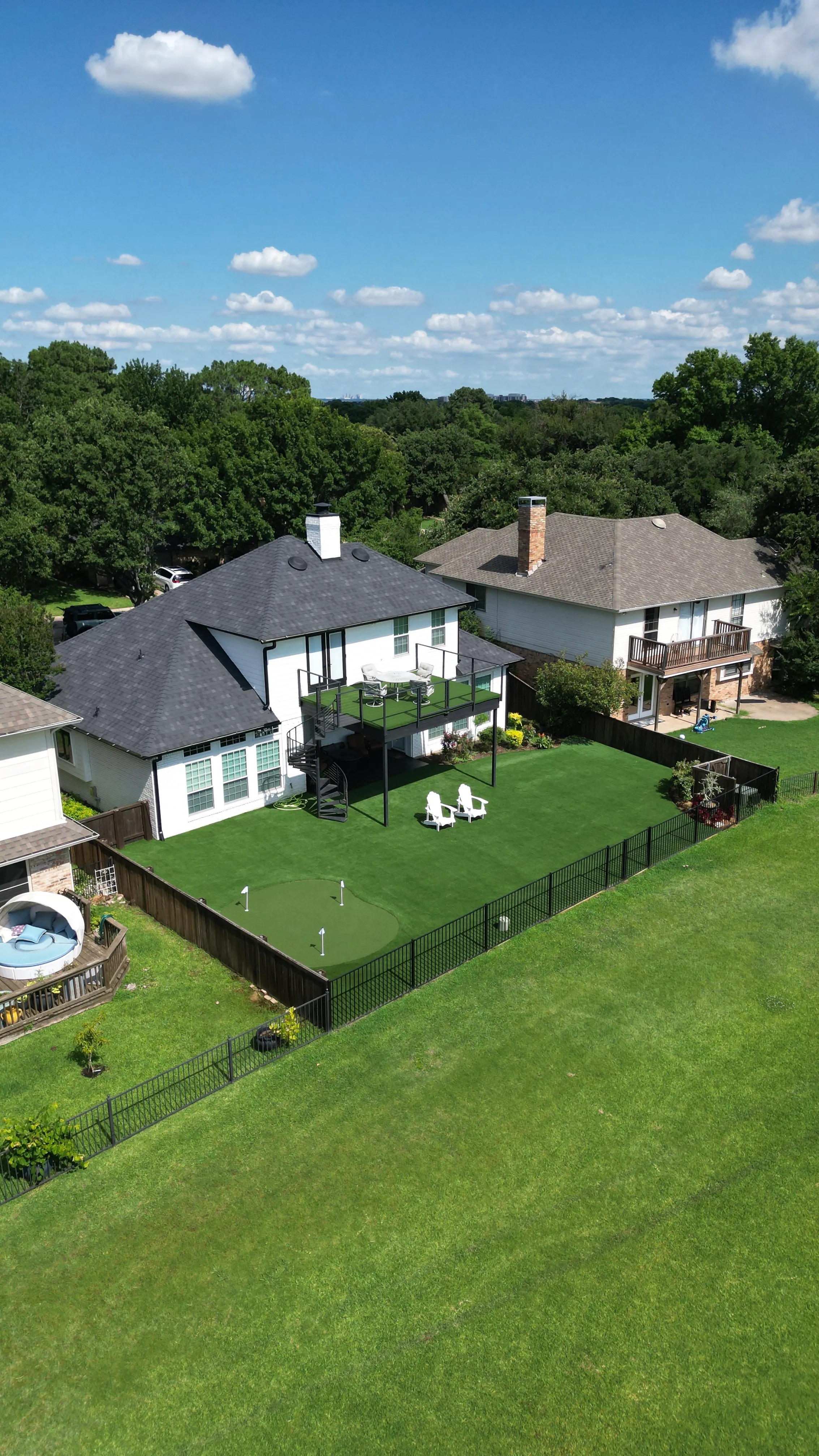 Aerial of a backyard with a rooftop turf deck over a spiral staircase, Adirondack chairs, and a putting green bordered by an iron fence