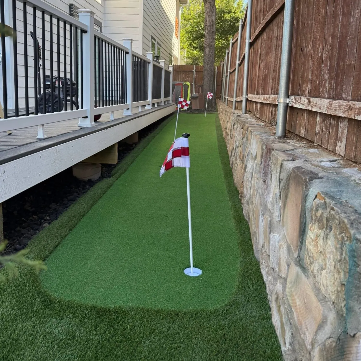 Narrow artificial putting green lane running beside a deck and stone-topped wood privacy fence in DFW