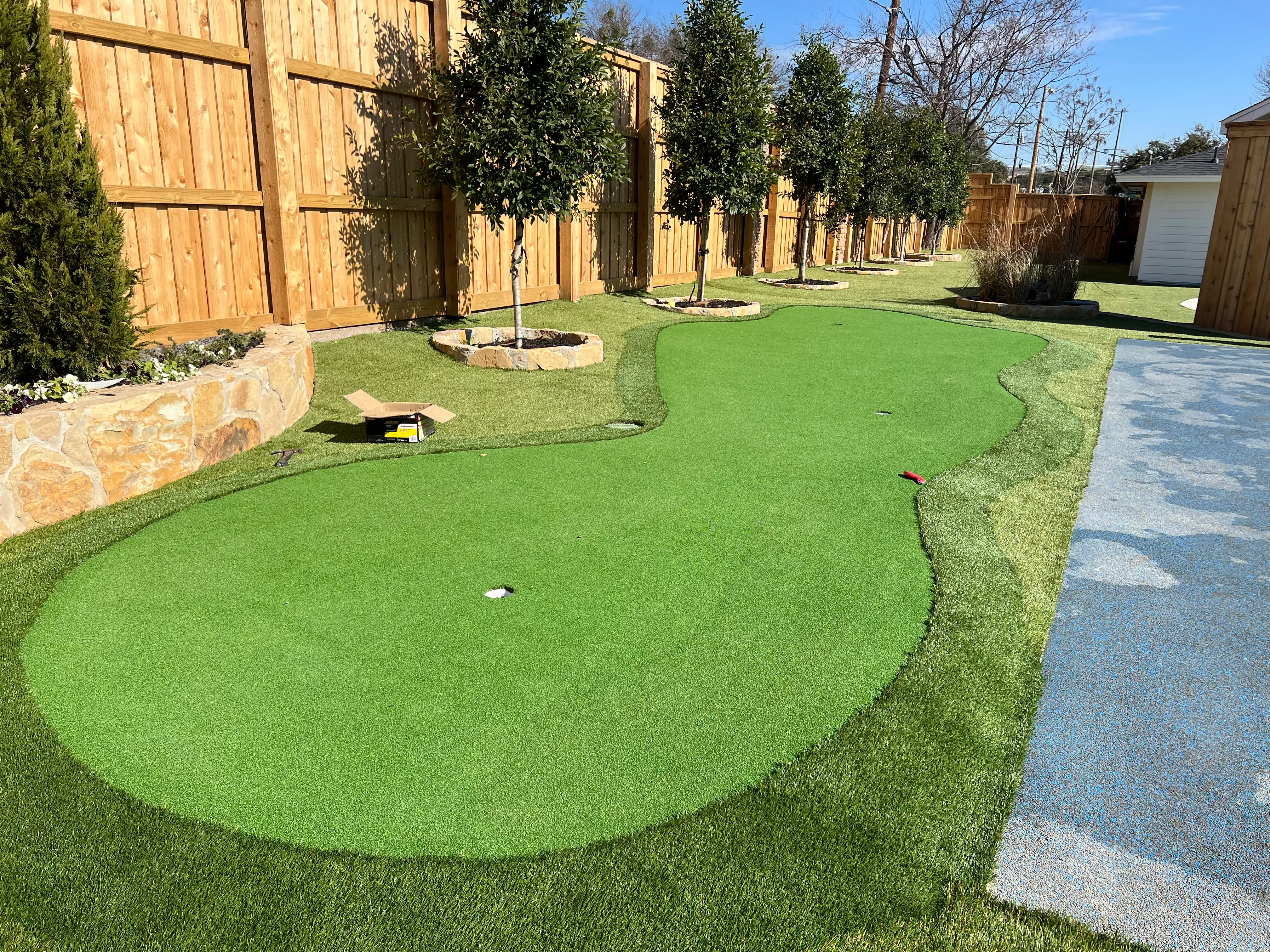Oval artificial putting green bordered by young ornamental trees in stone-ringed beds along a wood privacy fence in DFW
