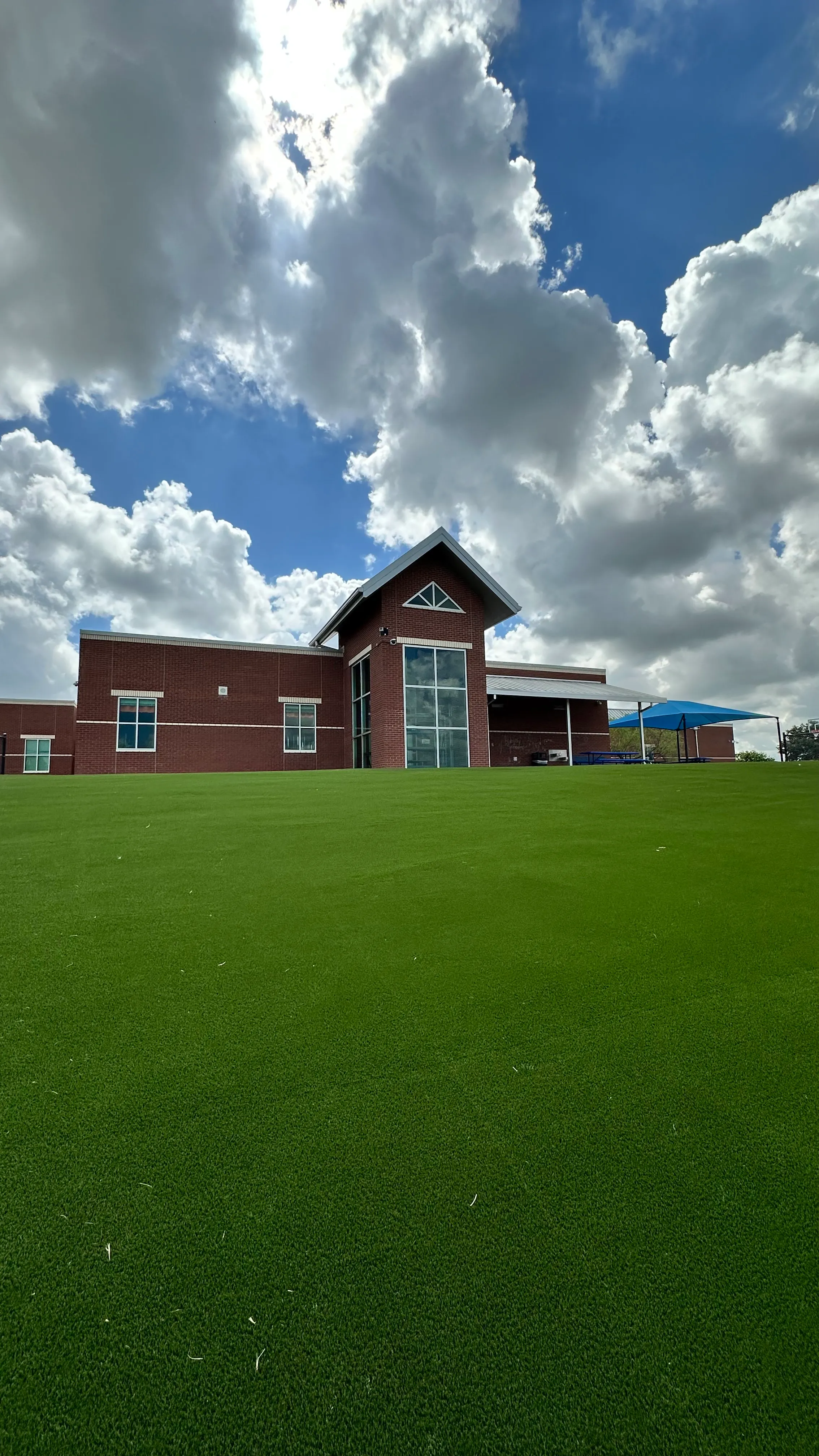 Low-angle view of graded artificial turf slope leading up to a DFW school building under dramatic clouds