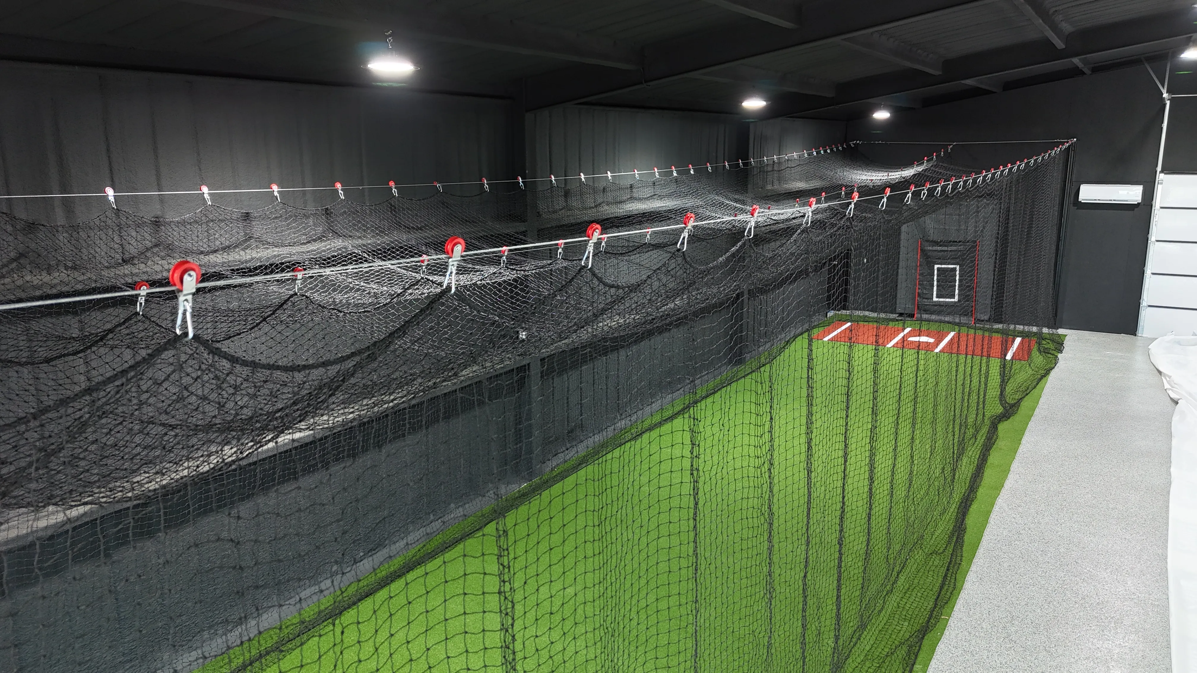 Angled interior view of an indoor batting cage building showing the netting rig and turf home plate area